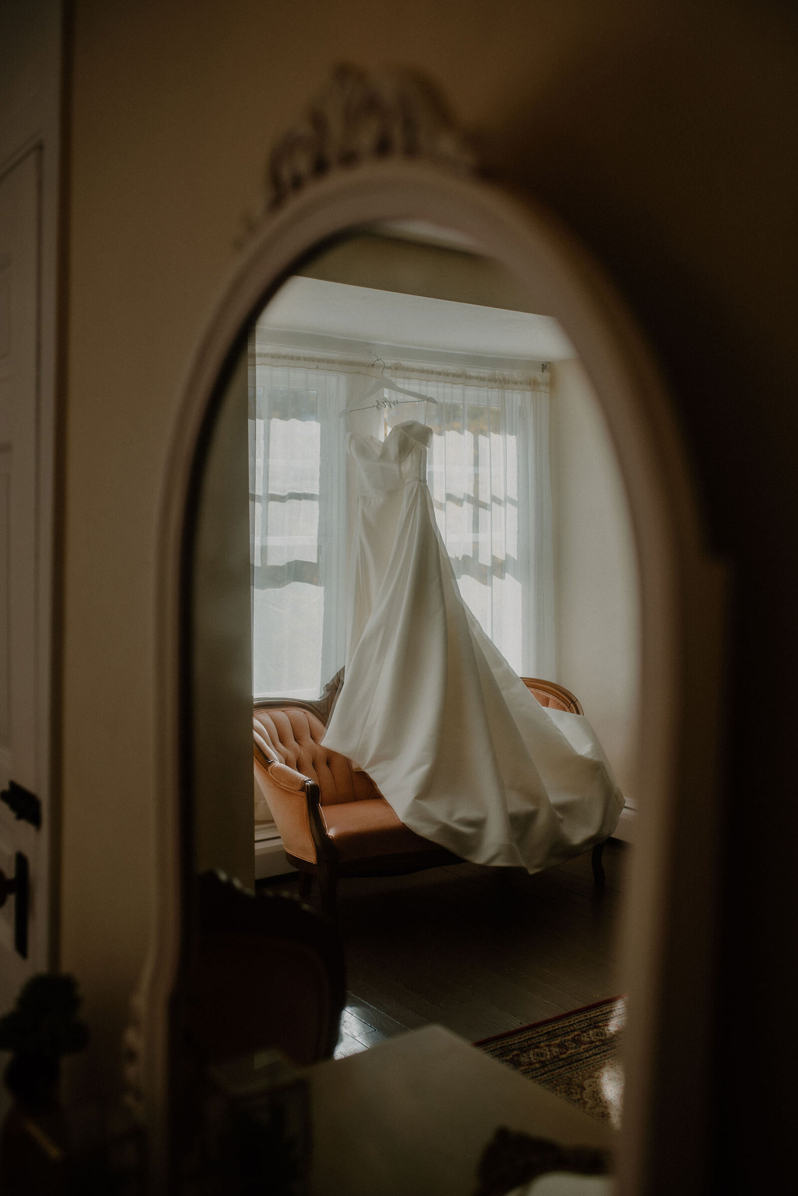 mirror reflection of wedding dress hanging in window over royal vintage couch