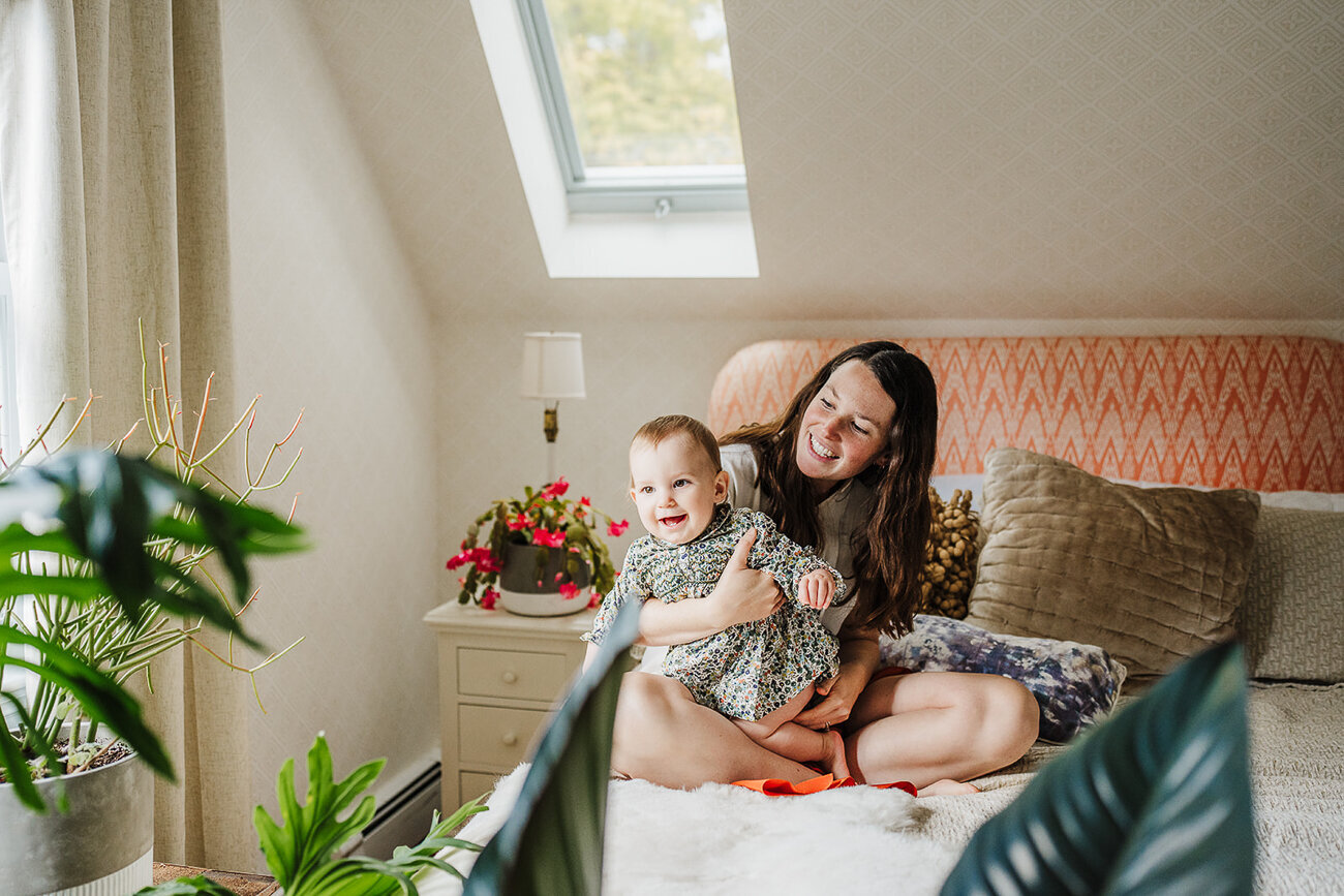 mother and toddler daughter laugh together on bed surrounded by plants