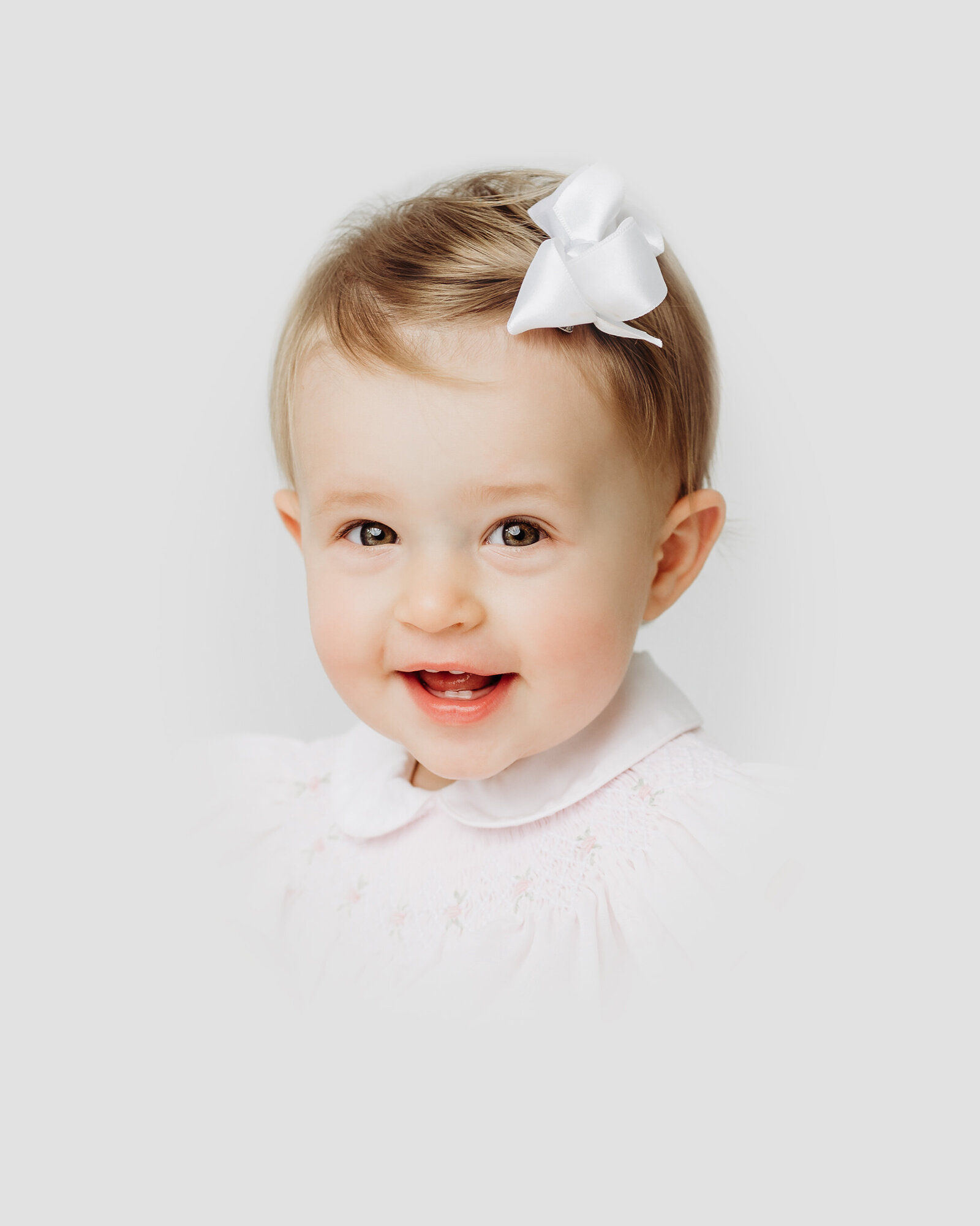 Little girl with white bow smiles at the camera during an heirloom portrait session in Raleigh NC