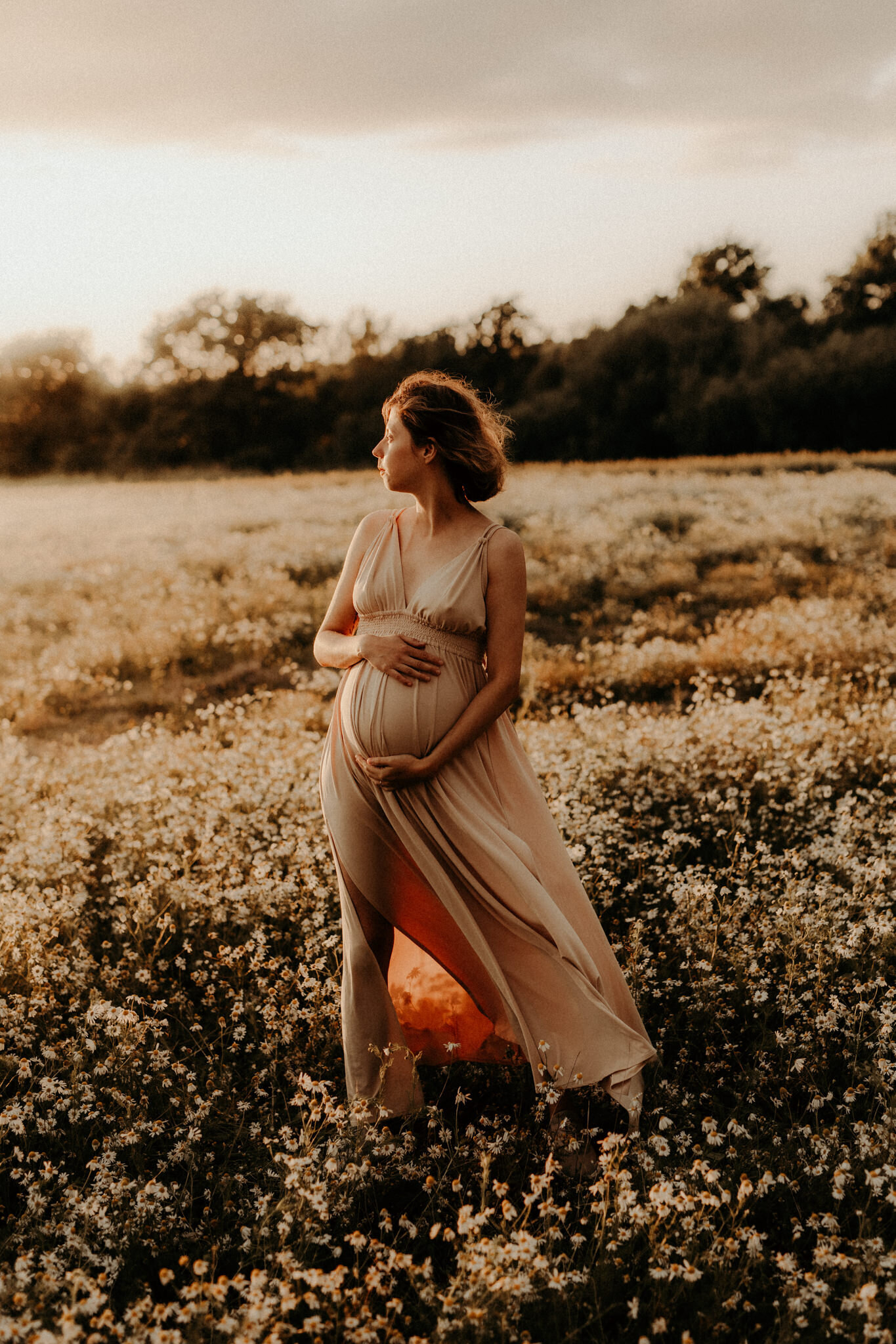 en gravid kvinna iklädd en beige gravidklänning står på ett fält med blommor