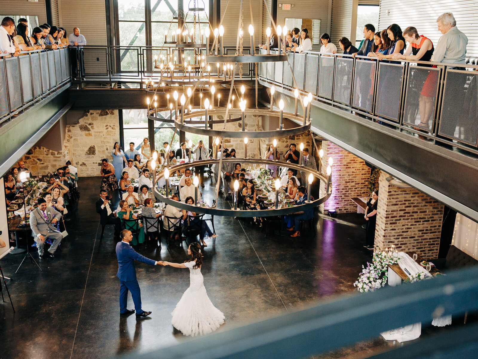 A bride and groom dance under large circular chandeliers in a modern, industrial-style venue as guests watch from tables and balconies—captured beautifully by a talented San Antonio Wedding Photographer.