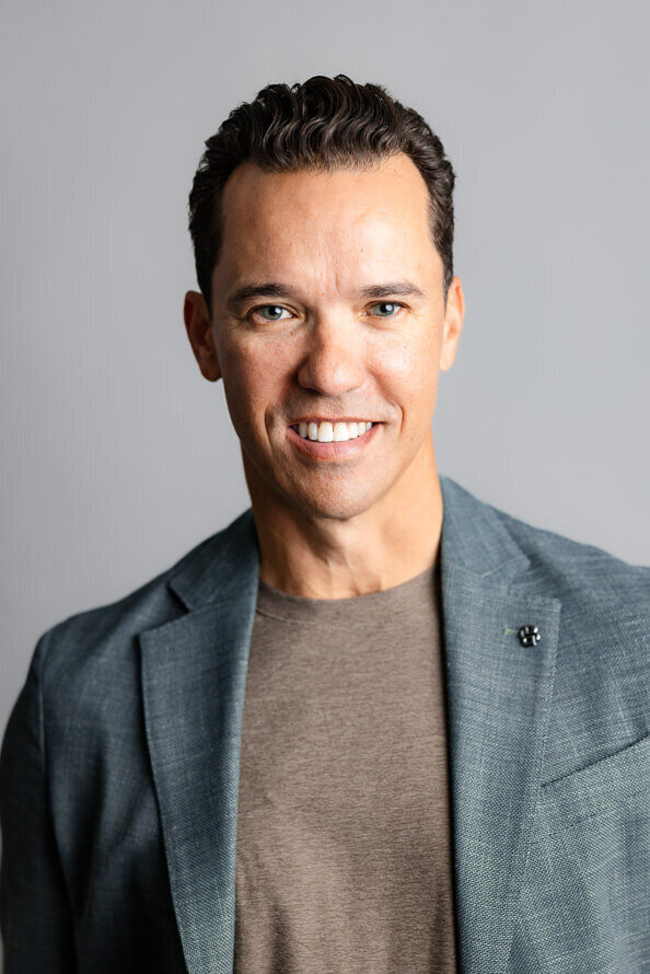Man in gray jacket smiles in front of gray backdrop.