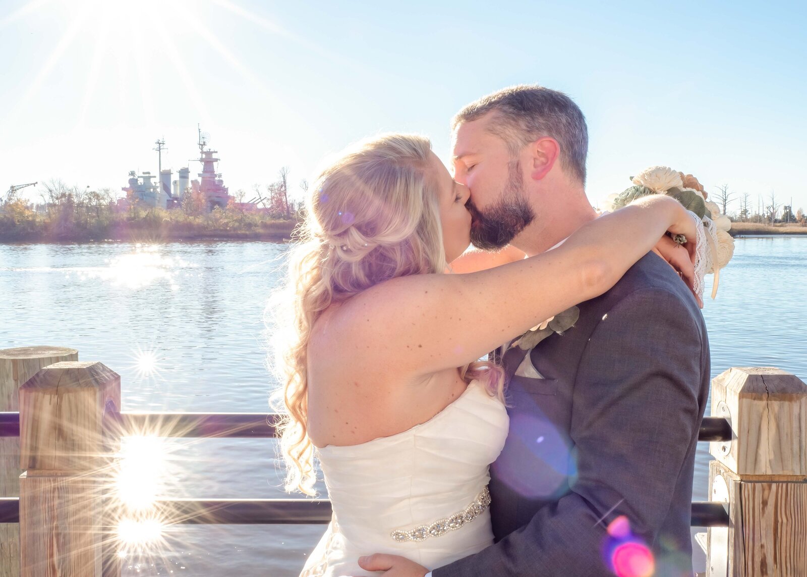 Romantic moment of the bride and groom sharing a kiss by the Cape Fear River in Wilmington, NC, with the USS North Carolina Battleship and sparkling sunlight in the background, showcasing timeless coastal beauty.