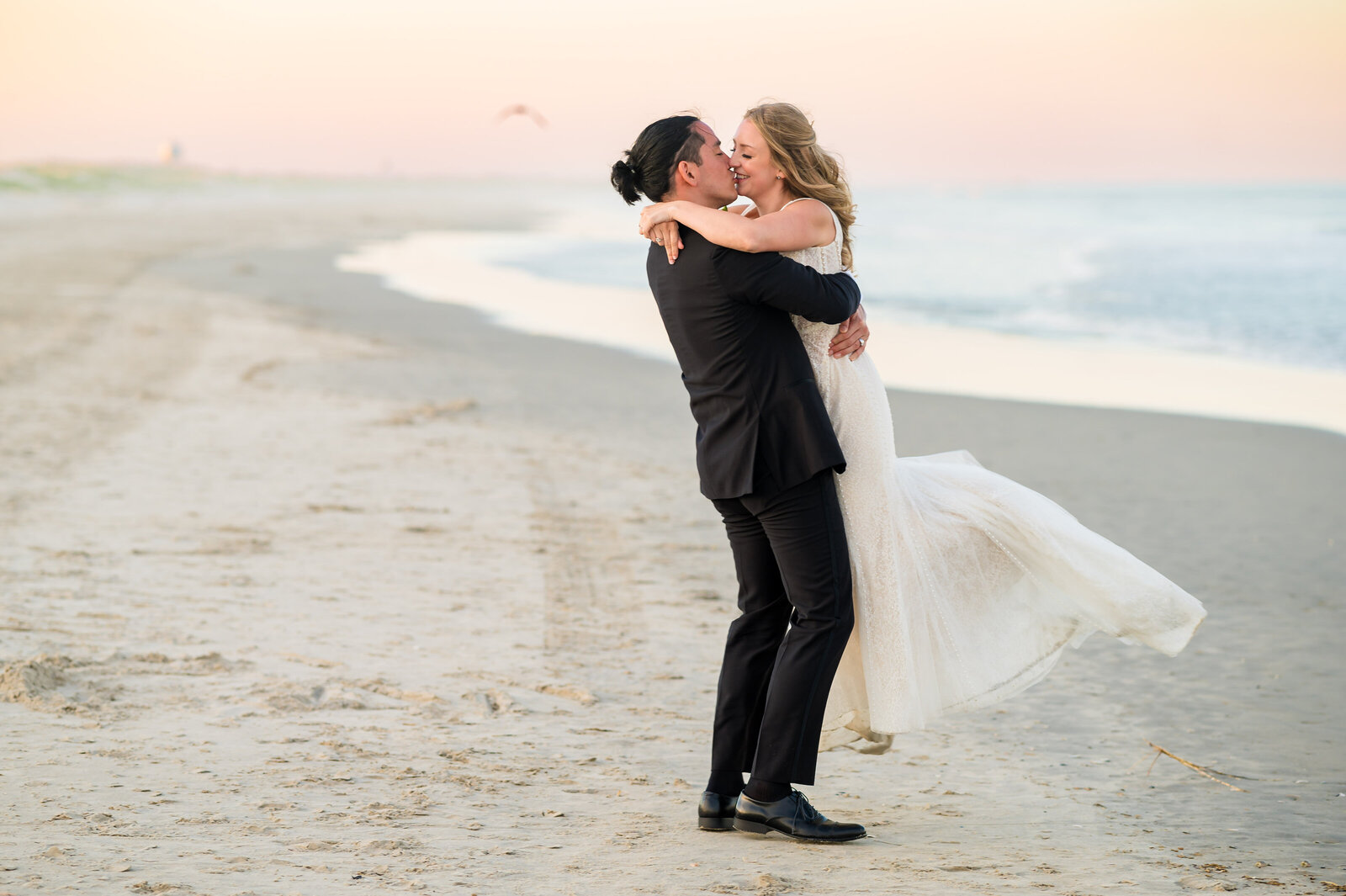 A couple is embracing and kissing on a sandy beach at sunset. The man is lifting the woman off the ground, and the ocean is visible in the background.