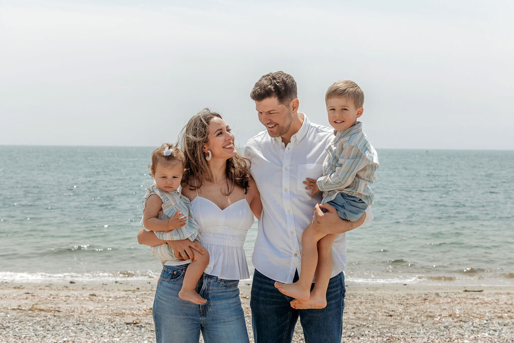 Joyful family of four laughing together on a Connecticut beach, captured by a Connecticut Family Photographer as parents hold their young children with ocean waves in the background
