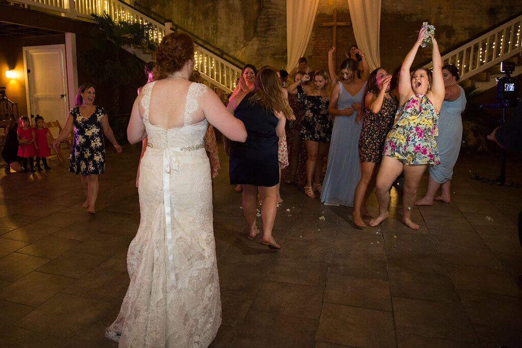 A bride in a lace gown faces a group of smiling women, some barefoot, as they celebrate catching her bouquet during a wedding reception, with a staircase and cross in the background.