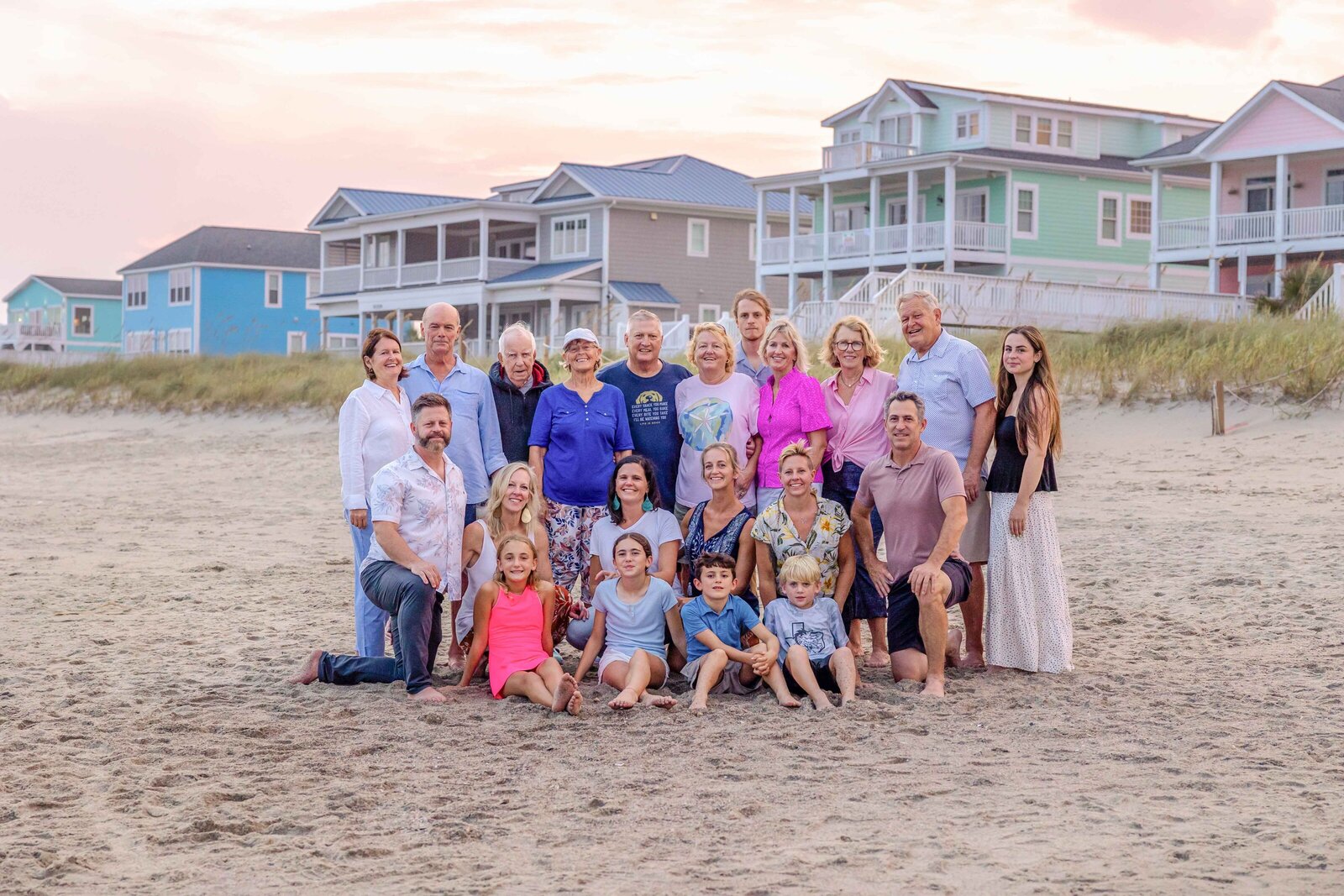 A large family poses on the beach with colorful beach houses behind them. Dressed in casual and colorful outfits, they enjoy a lively and joyful scene, ideal for destination family photographer NC, capturing vibrant family connections by the sea.