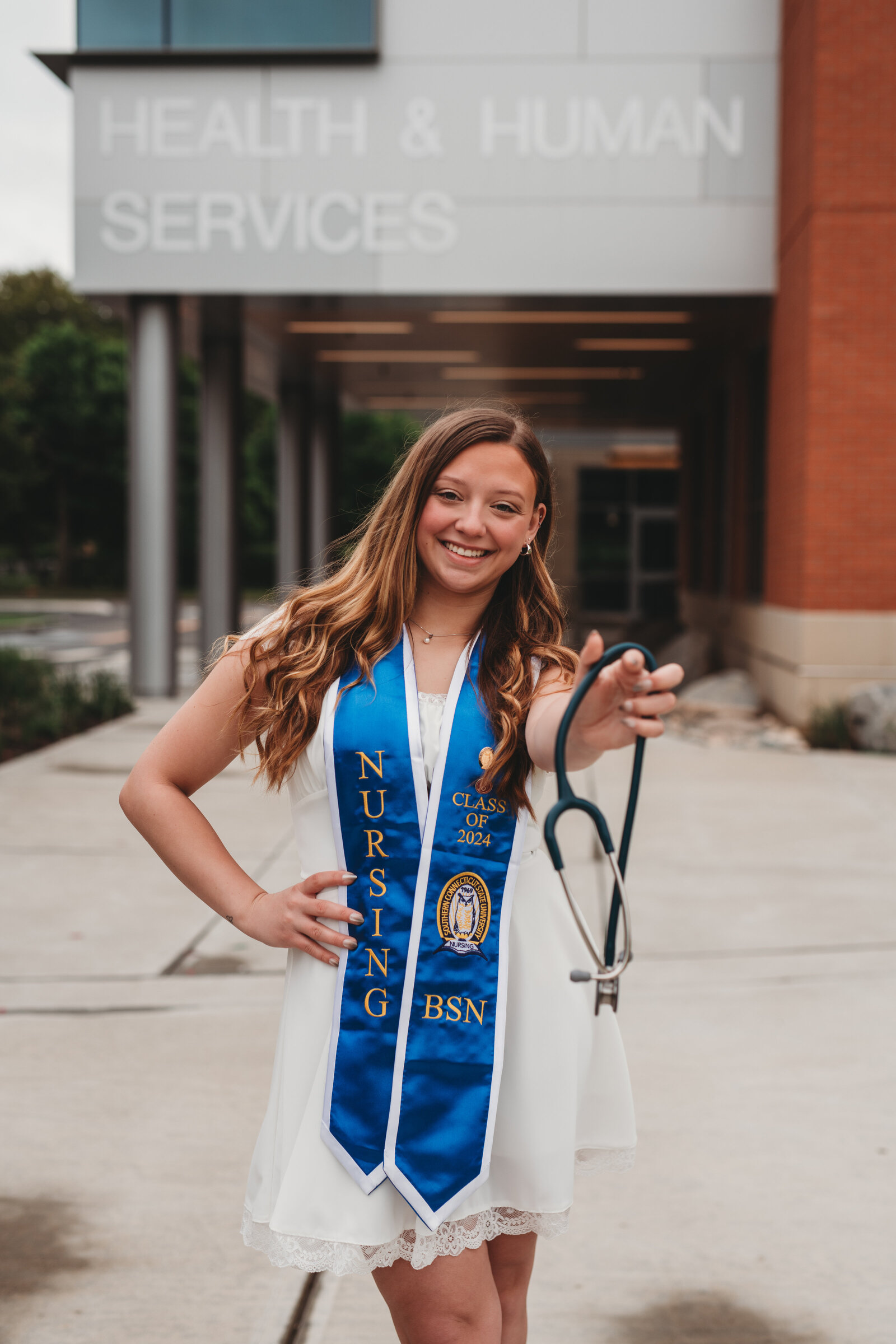 A proud Southern Connecticut State University nursing graduate in a white dress and blue BSN stole poses in front of the Health & Human Services building, holding a stethoscope. Captured during a Connecticut nursing graduation photography session by Nicole Mele Photography, highlighting achievement and dedication.