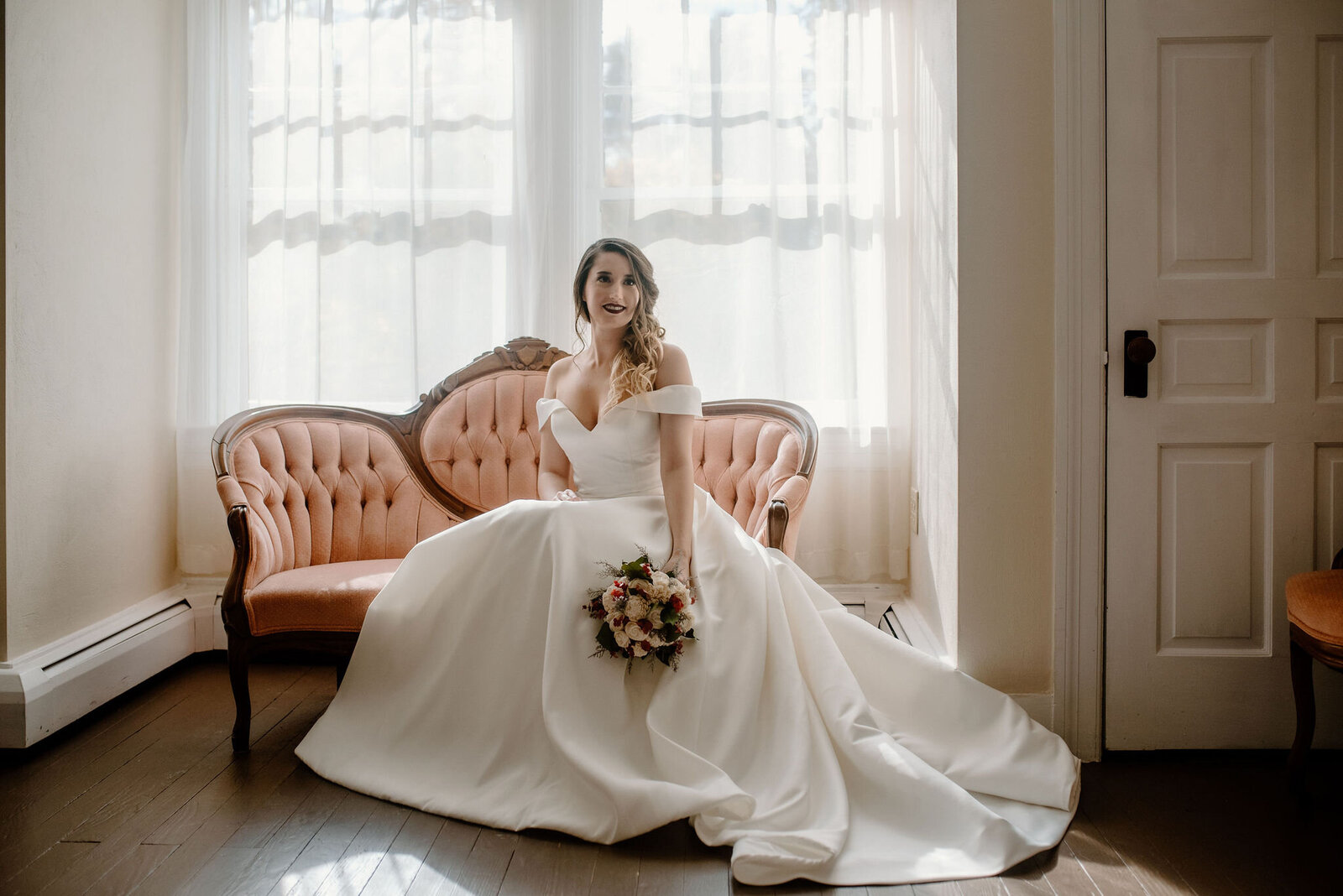 bride sitting on regal, vintage pink couch in wedding dress with white and red flower bouquet