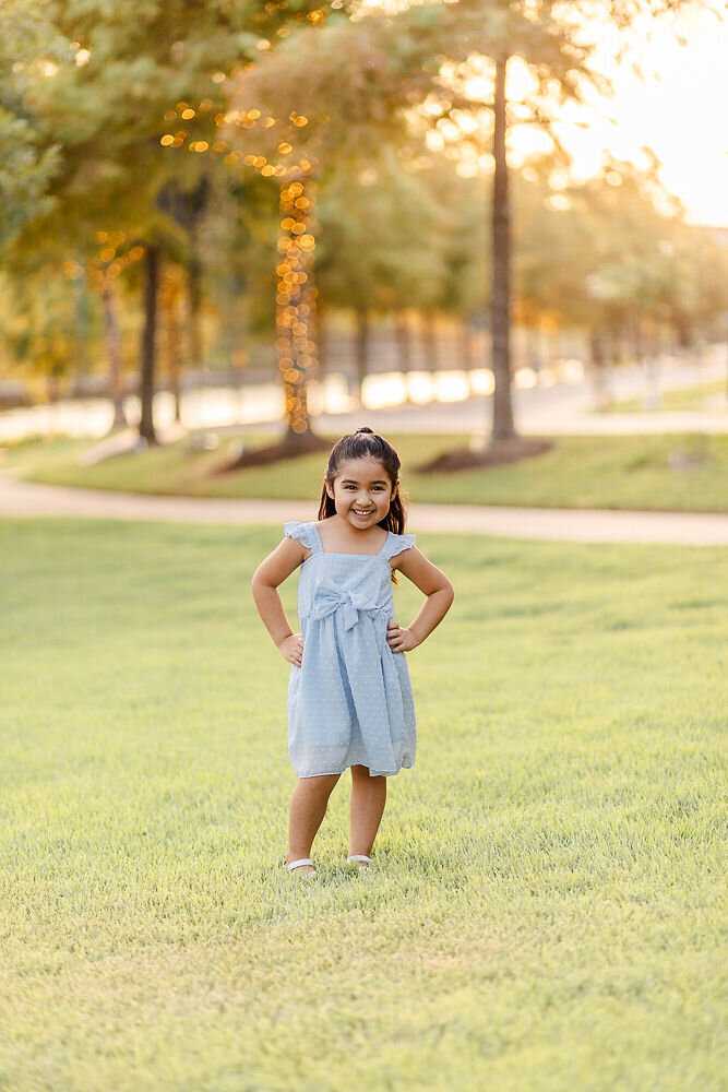 A young girl stands confidently on a grassy lawn during golden hour, smiling brightly with her hands on her hips. She wears a light blue dress with a large bow on the front and white sandals. Behind her, tall trees with soft, glowing string lights create a warm, dreamy backdrop along a winding pathway.