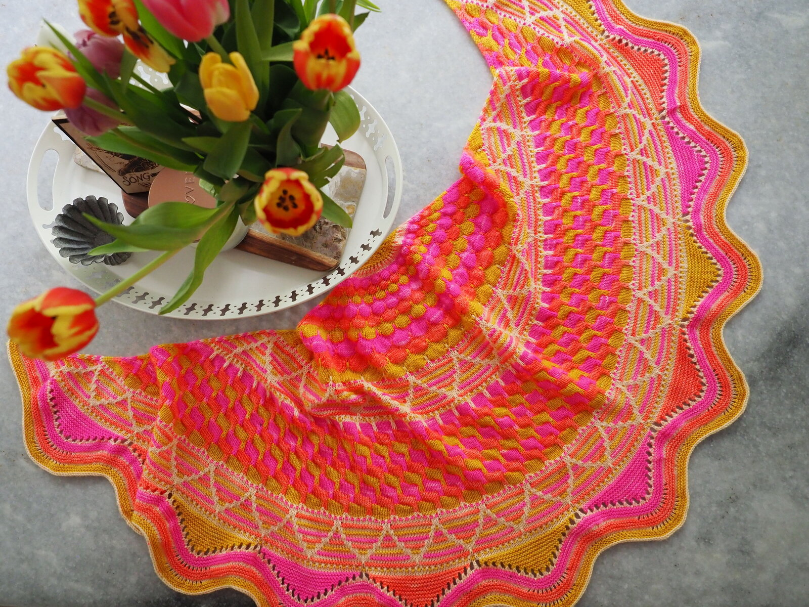 Top view of a hand knit orange, pink and yellow shawl with a vase of tulips next to it.