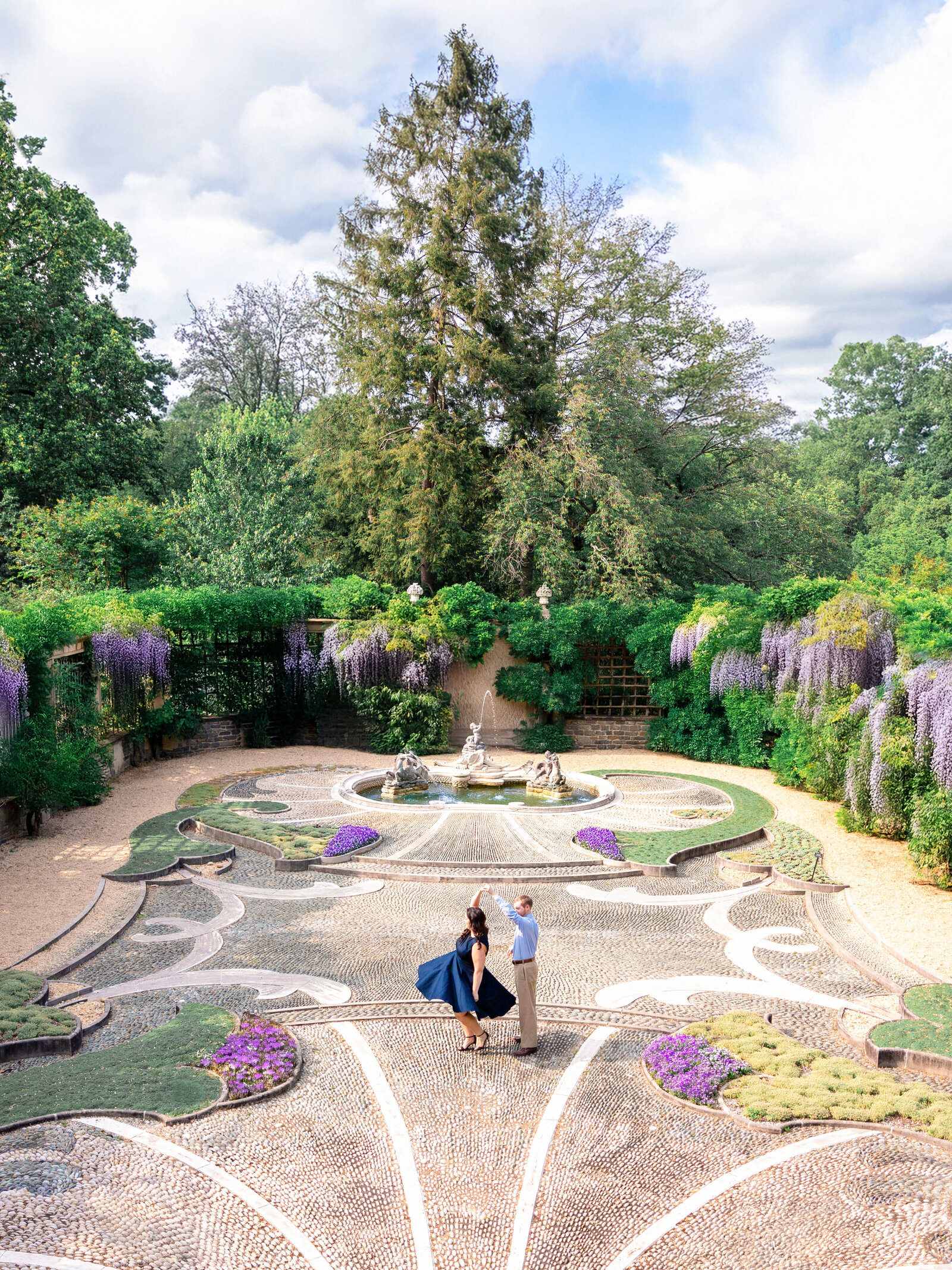 A couple dances in a beautifully landscaped garden with intricate stone pathways and purple flower arrangements. Tall trees and lush greenery surround them under a partly cloudy sky.