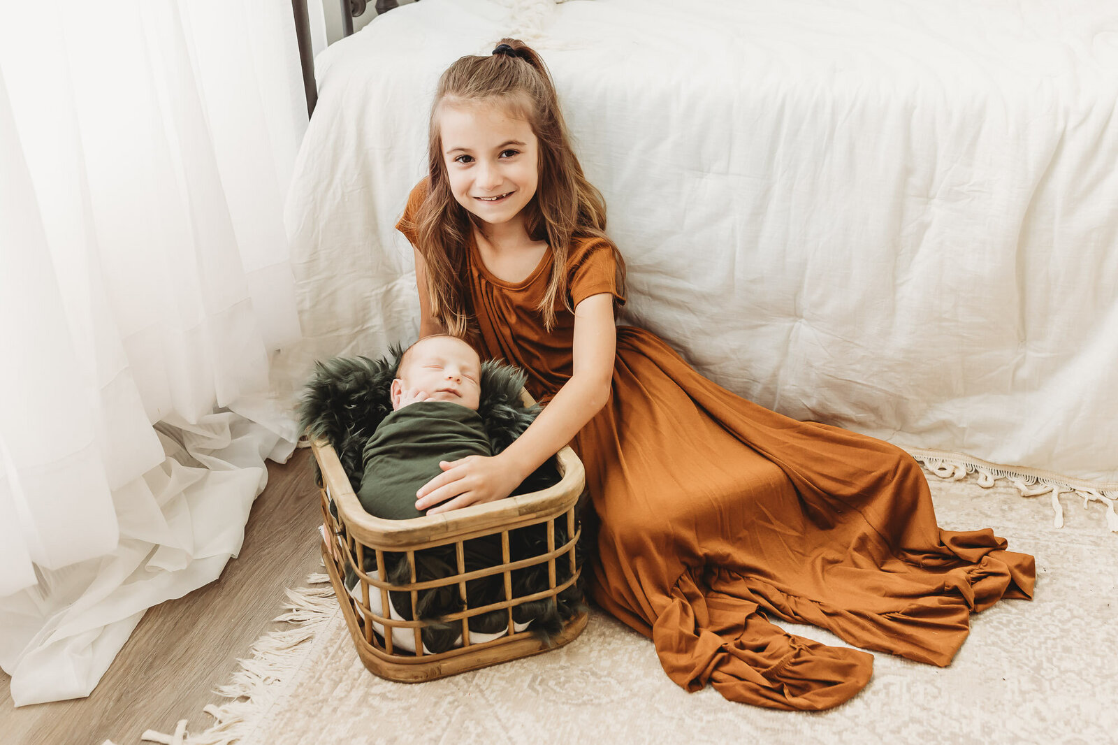 Big sibling in a copper colored dress laying next to baby brother who is wrapped in an olive green swaddle in a basket