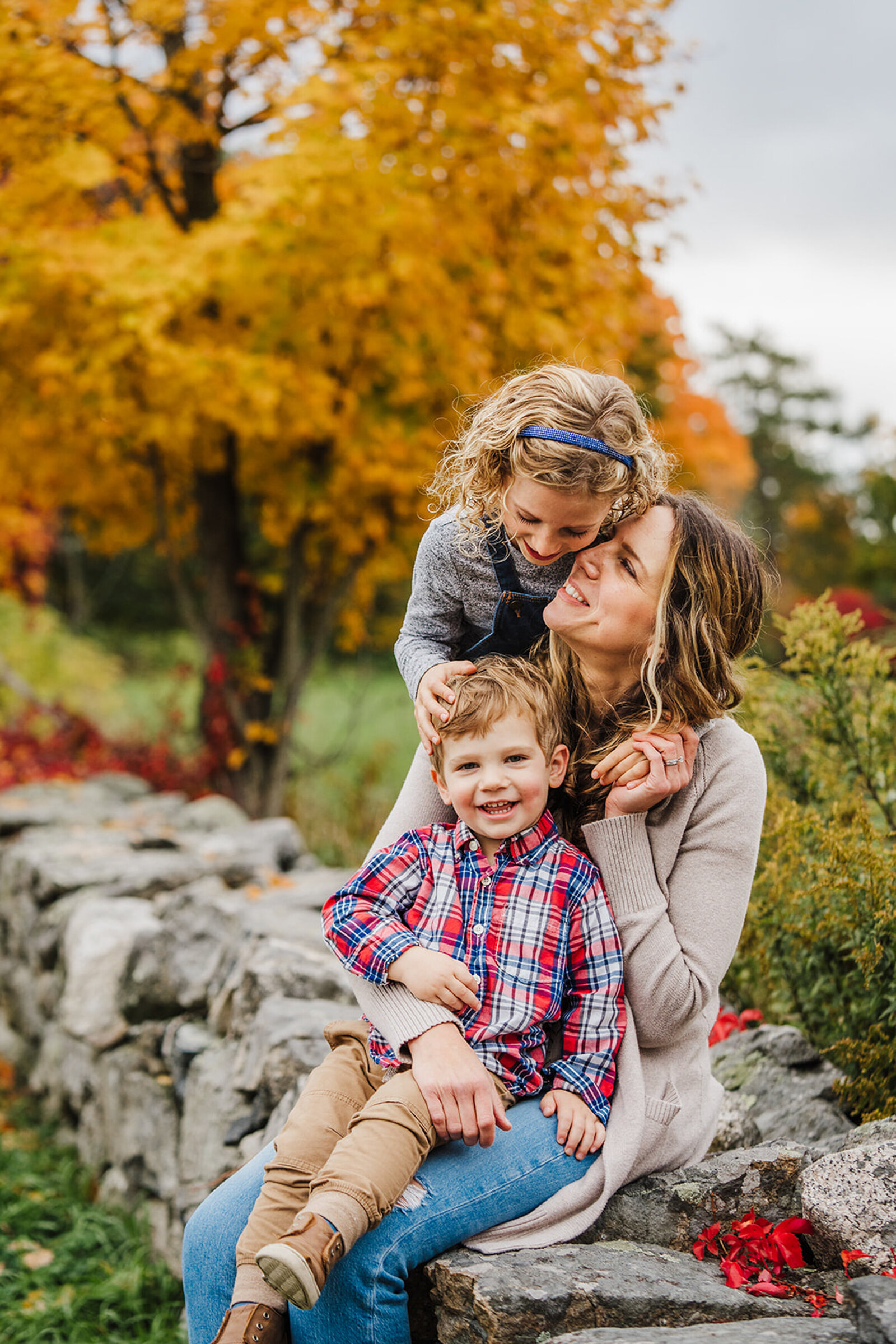 mom snuggles two kids on a wall with autumn leaves