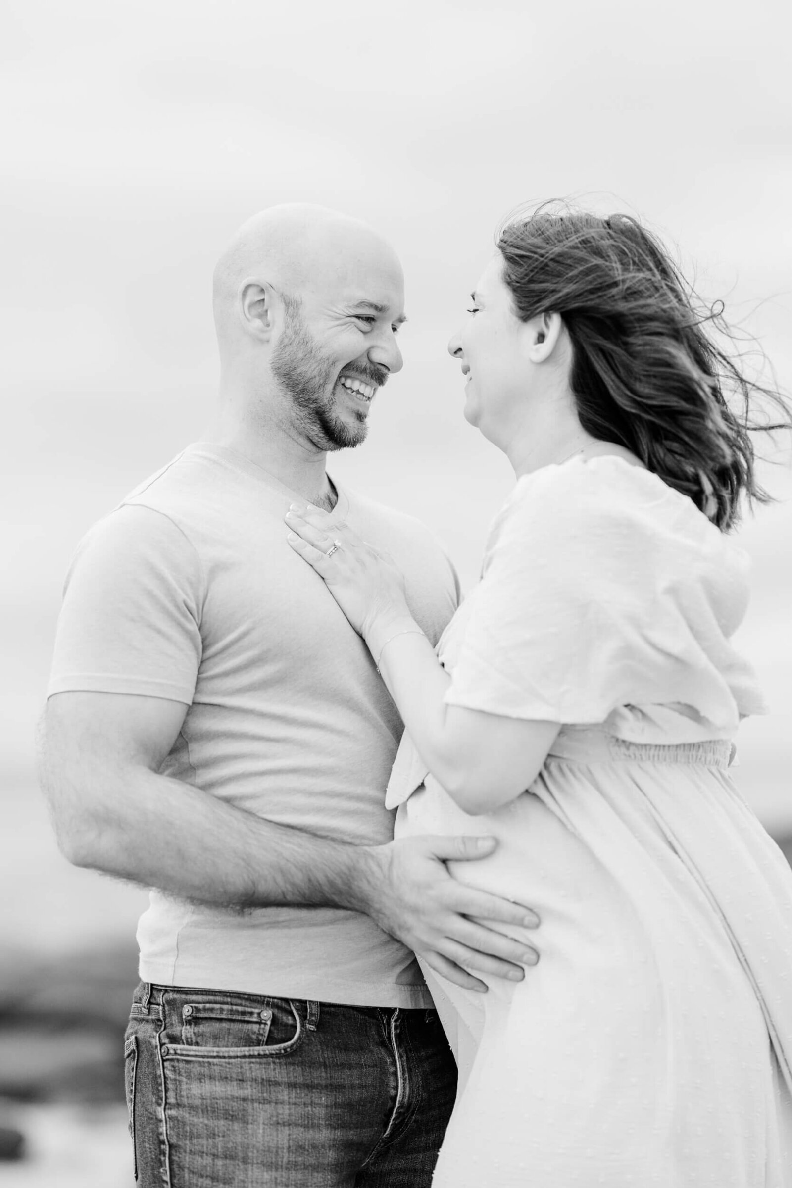 Black and white, man embraces his pregnant wife's bump and laughs as the wind blows her hair and dress