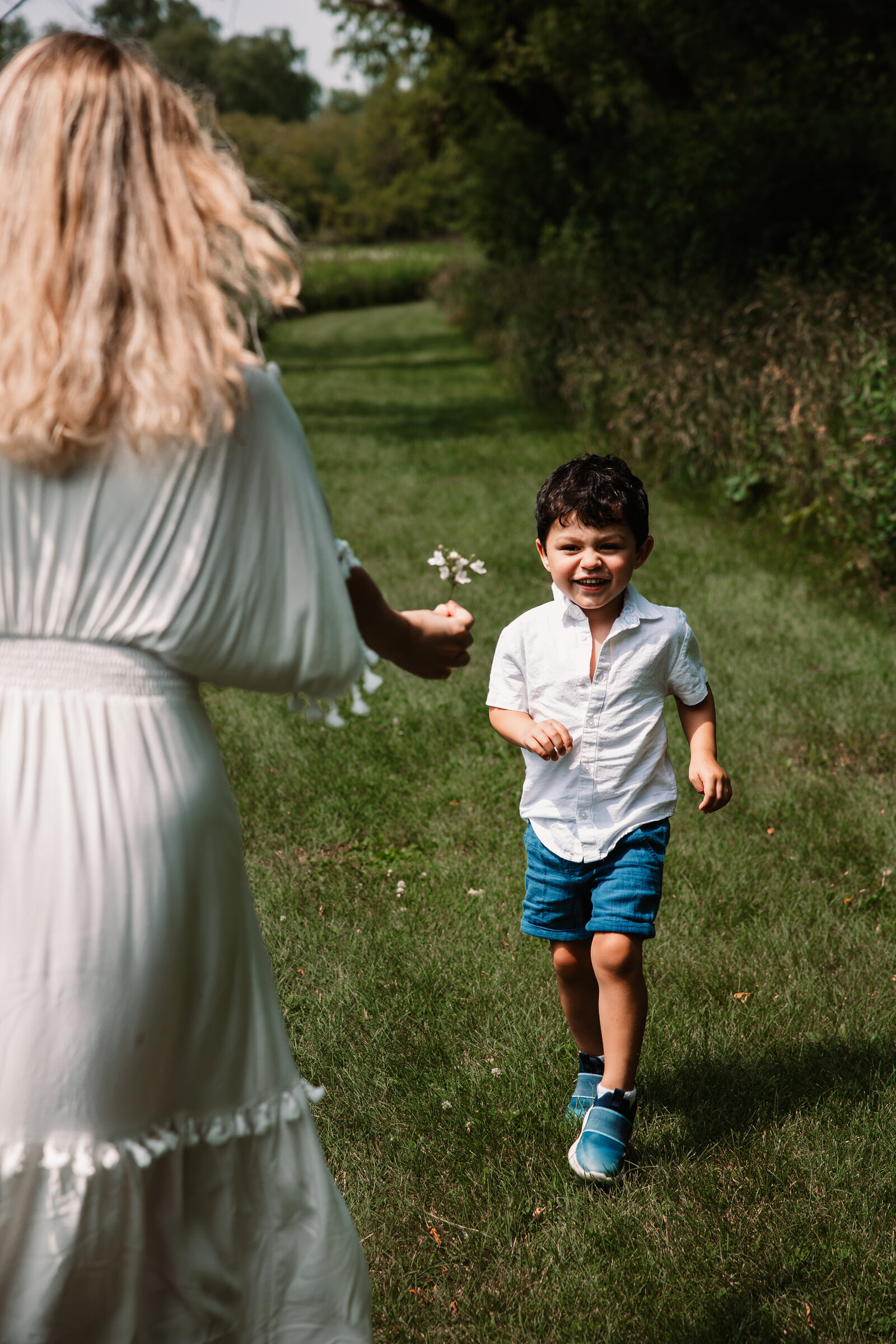 A mom in a white dress holds flowers as her toddler son run towards her in a park trail captured by a Milwaukee Family Photographer