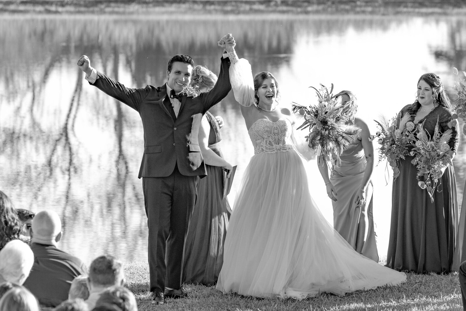 The bride and groom celebrate joyfully with hands raised after their lakeside wedding ceremony in Boone, NC, surrounded by a serene autumn backdrop.