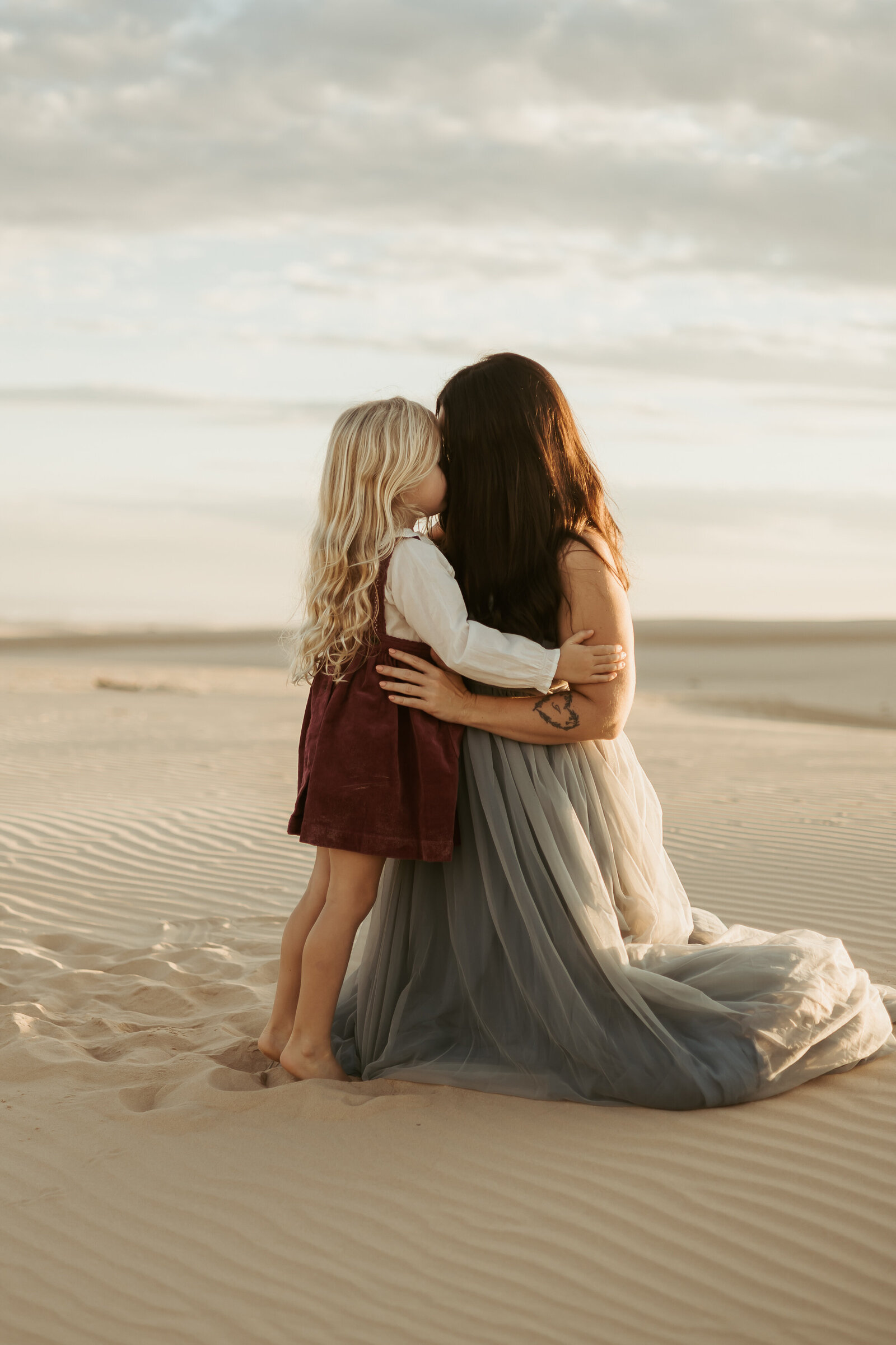 A heartfelt moment captured of a mother and daughter sharing a cuddle as the sun sets behind them on the sand dunes.