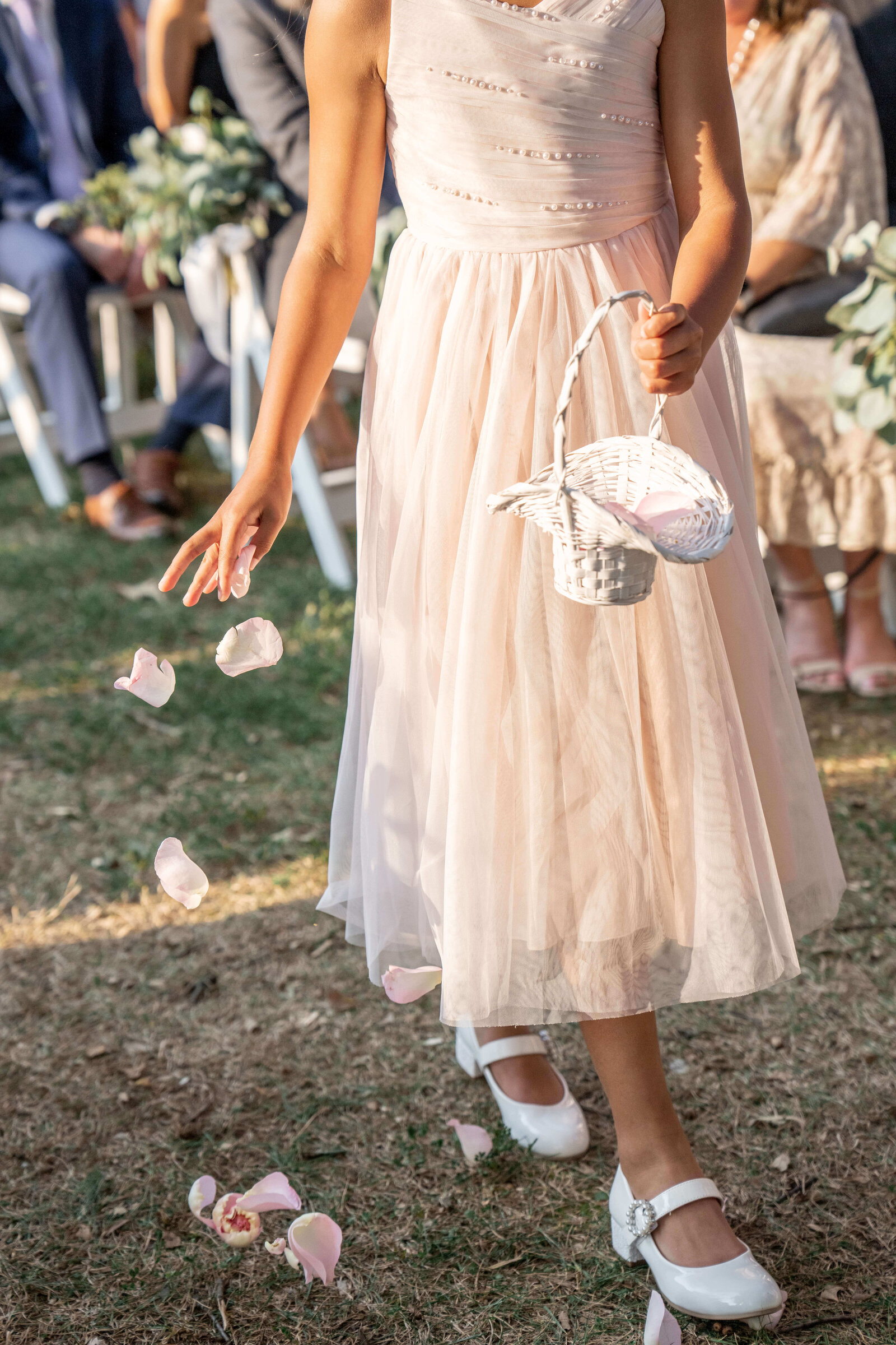 A young girl in a light pink dress scatters rose petals from a white basket as she walks on grass during a wedding ceremony. She wears white shoes, and guests are seated in the background.