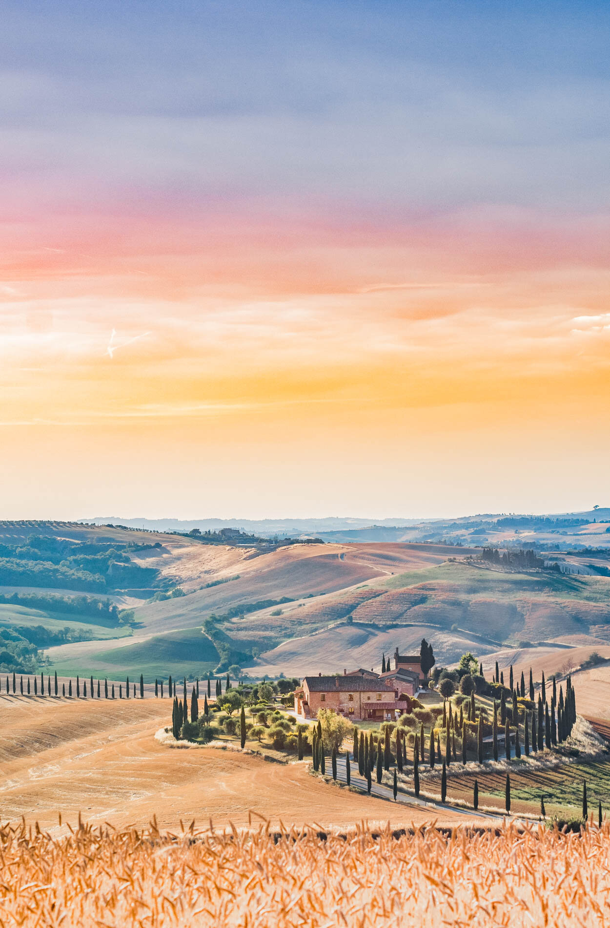 The rolling hills of Tuscany in Italy featuring a beautiful stone house in the foreground with cypress trees all around.