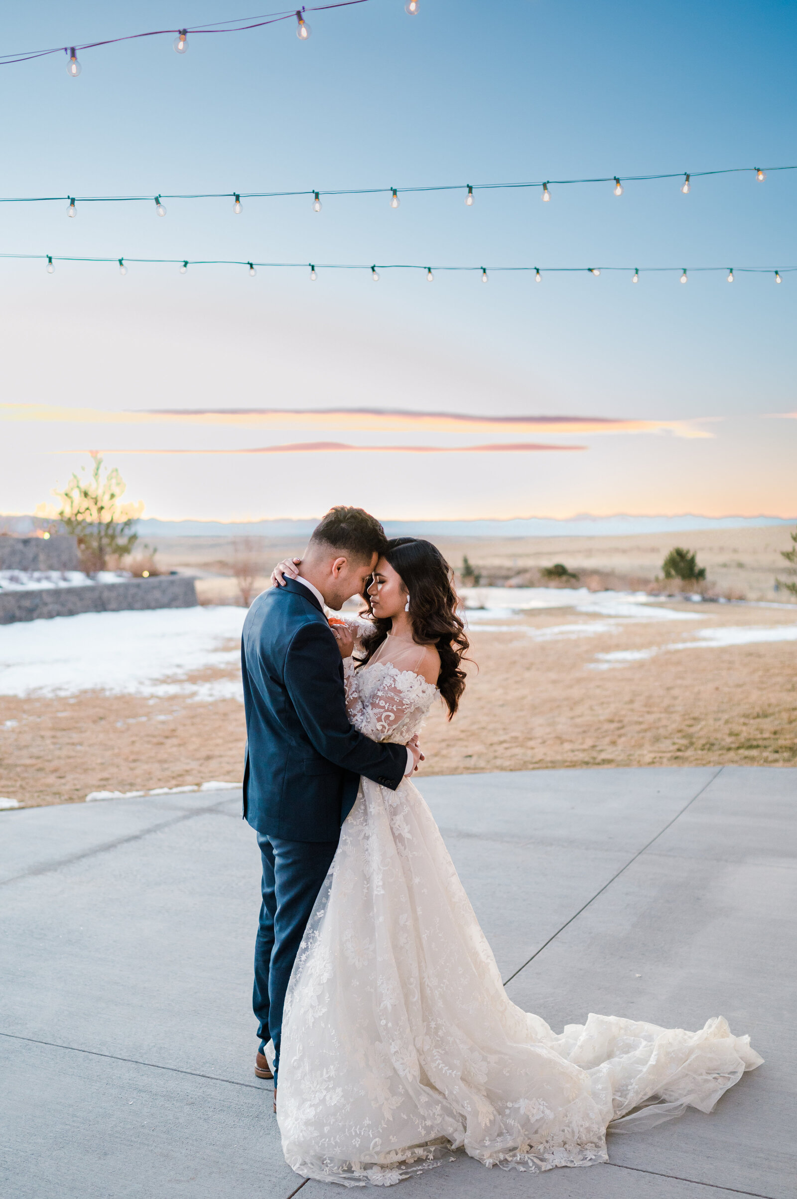 Bride in gorgeous strapless dress and Groom in handsome suit embrace during their first dance at sunset on patio
