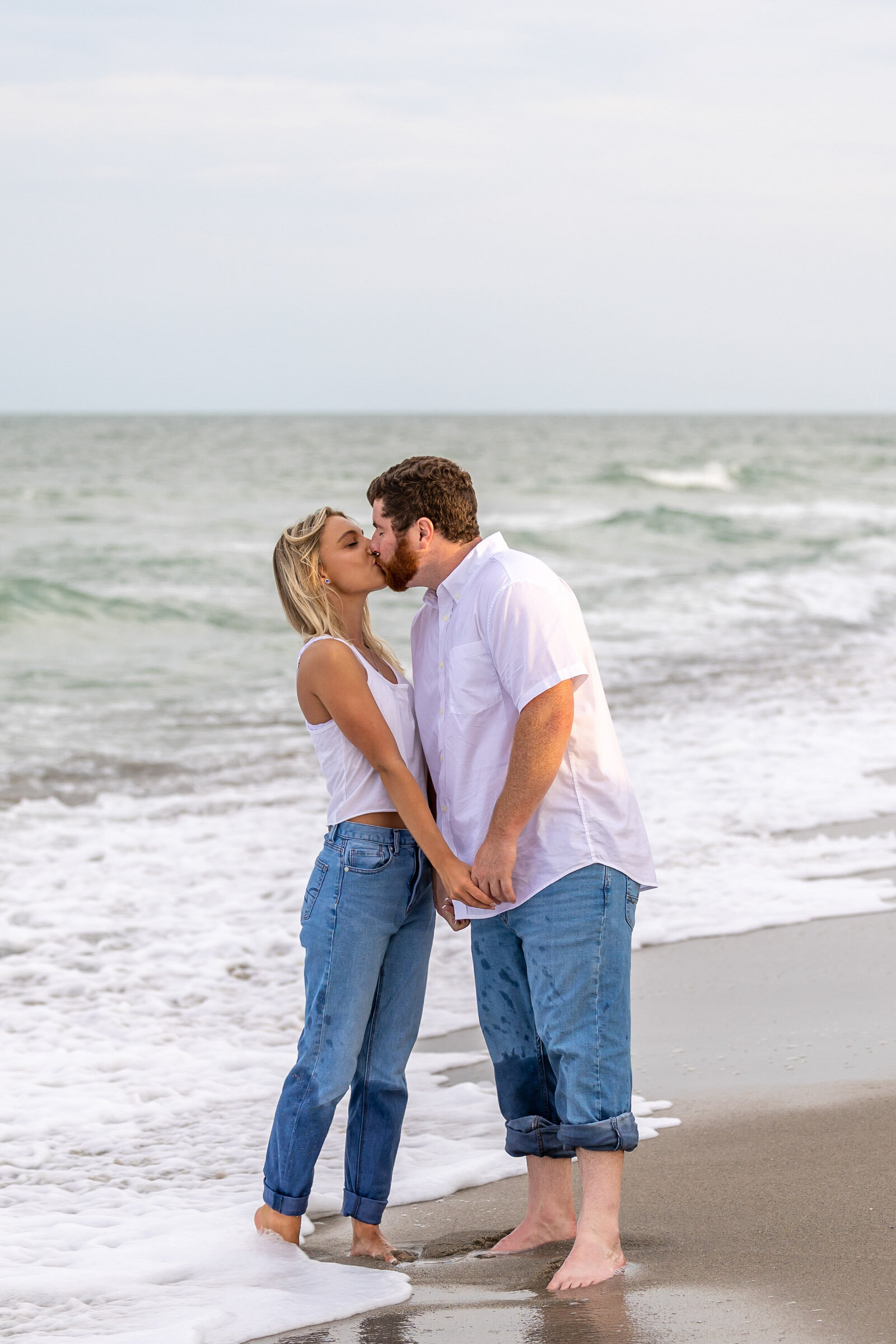 A couple sharing a romantic kiss on the beach in Wilmington, NC, with waves gently lapping at their feet. This image reflects the essence of authentic wedding photography and showcases the beauty of simultaneous photo and video services for couples.