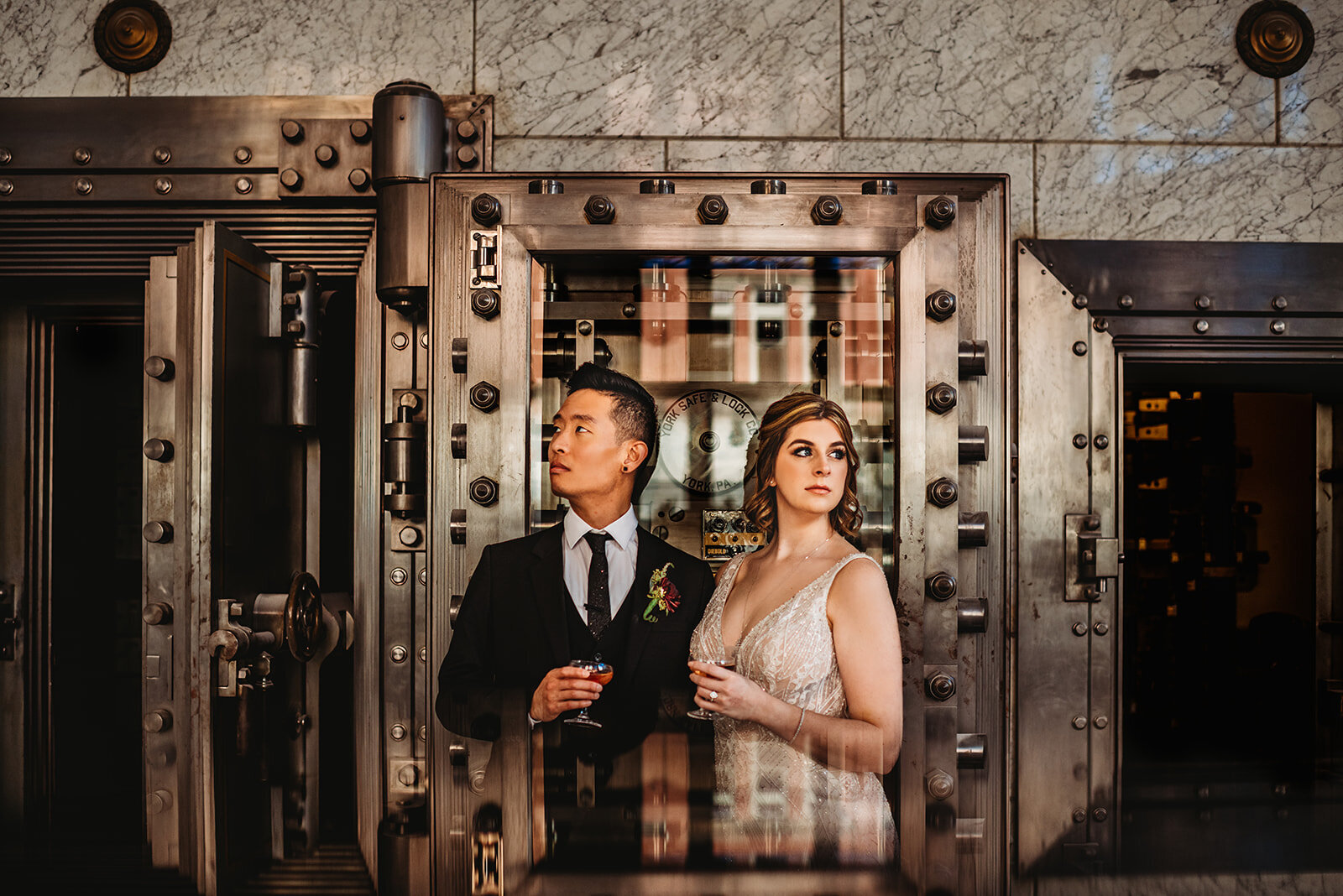 Baltimore photographers captures wedding at a bank with bride and groom holding cocktails while standing in front of a vault