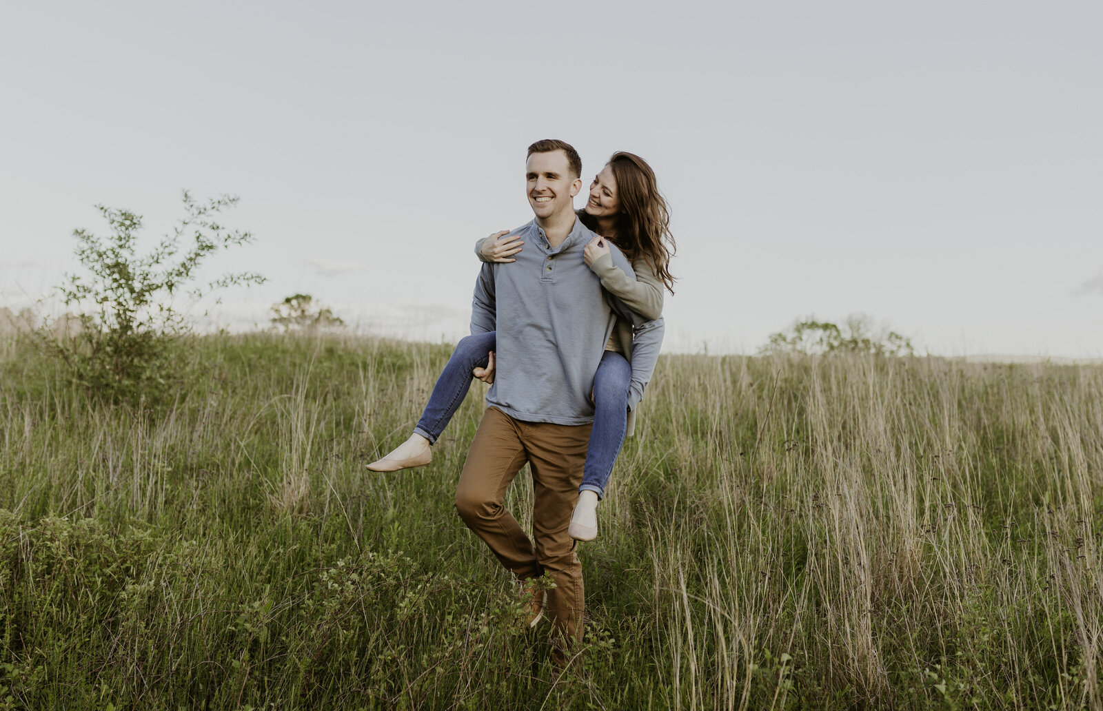 A man piggybacks a woman in a field of grass at sunset