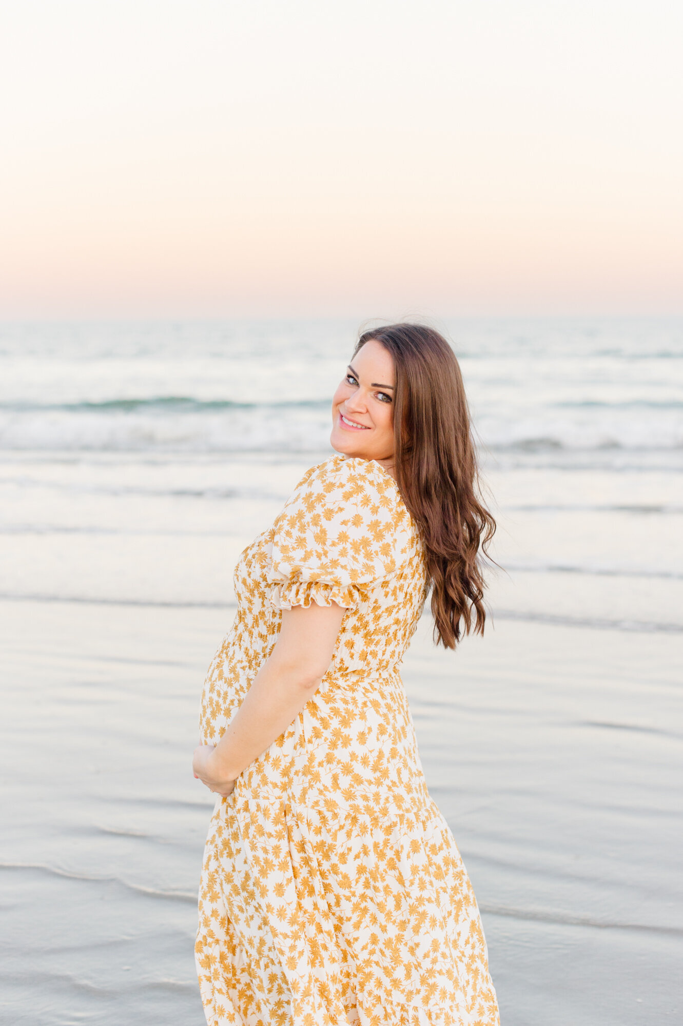 Pregnant mother wearing yellow floral looking over her shoulder smiling at the camera during her maternity photo session