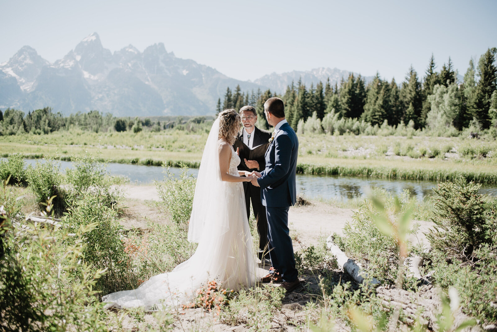 summer wedding captured by jackson hole wedding  photographer with bride and groom holding hands and listening to their officiant in front of a creek with the grand tetons in the distance