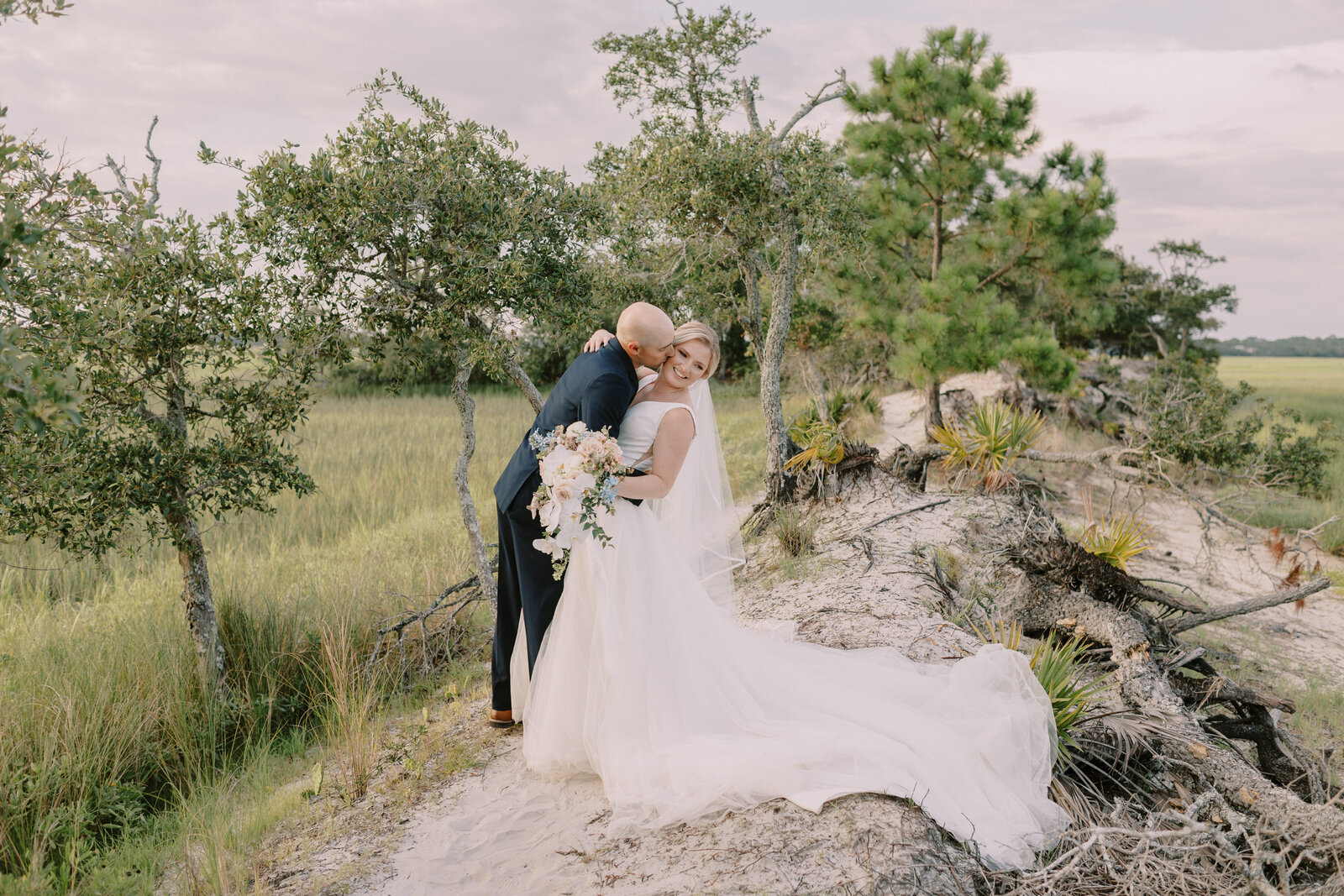 Image by Savannah wedding photography image of groom in navy tuxedo dipping bride holding a cascading bouquet of pastel florals while kissing her cheek. Her long train hangs over the side of a peak overlooking the summertime marsh with lush greens overcast sky.