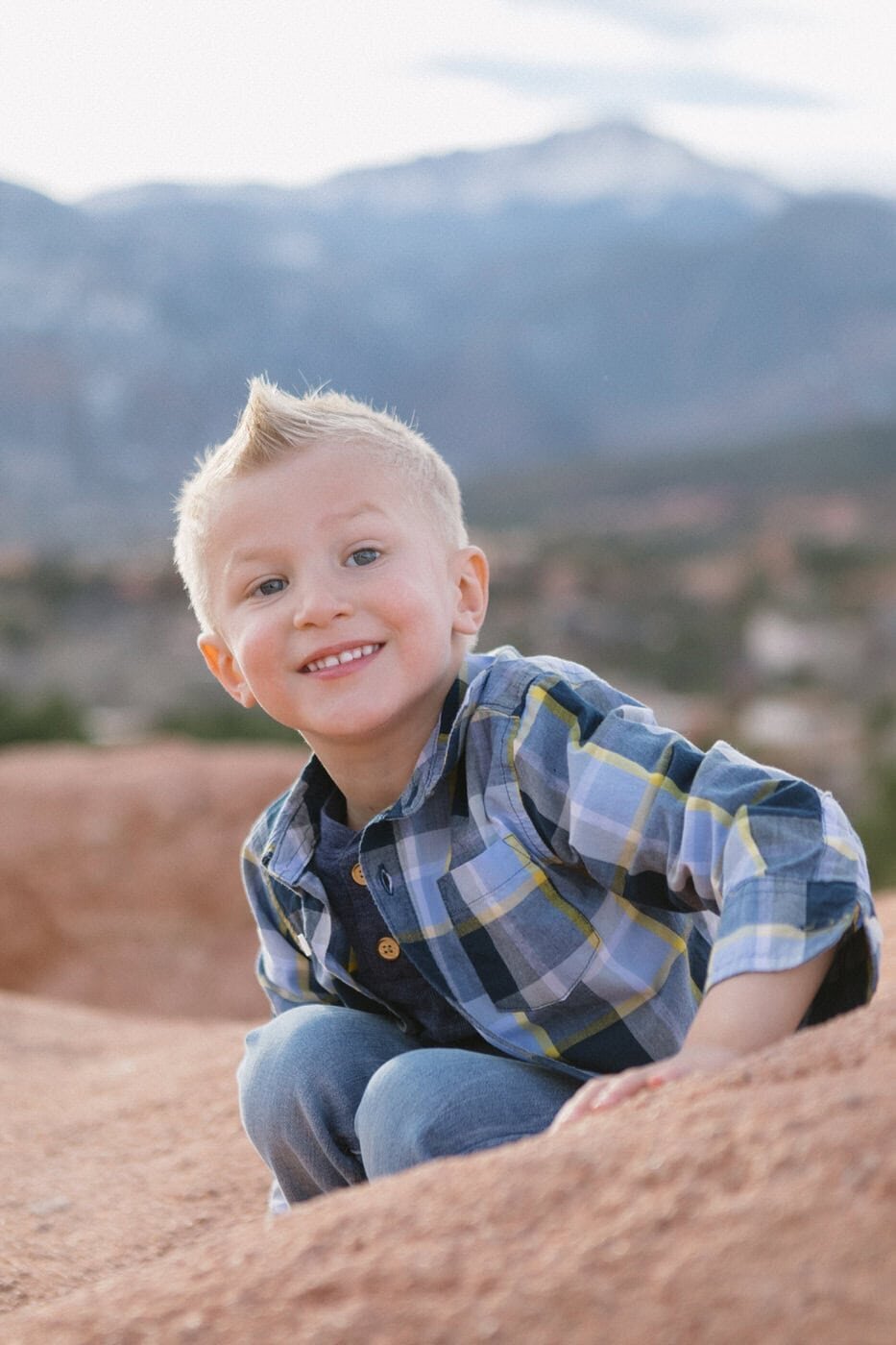 A boy crouches on a red rock and smiles towards the camera.