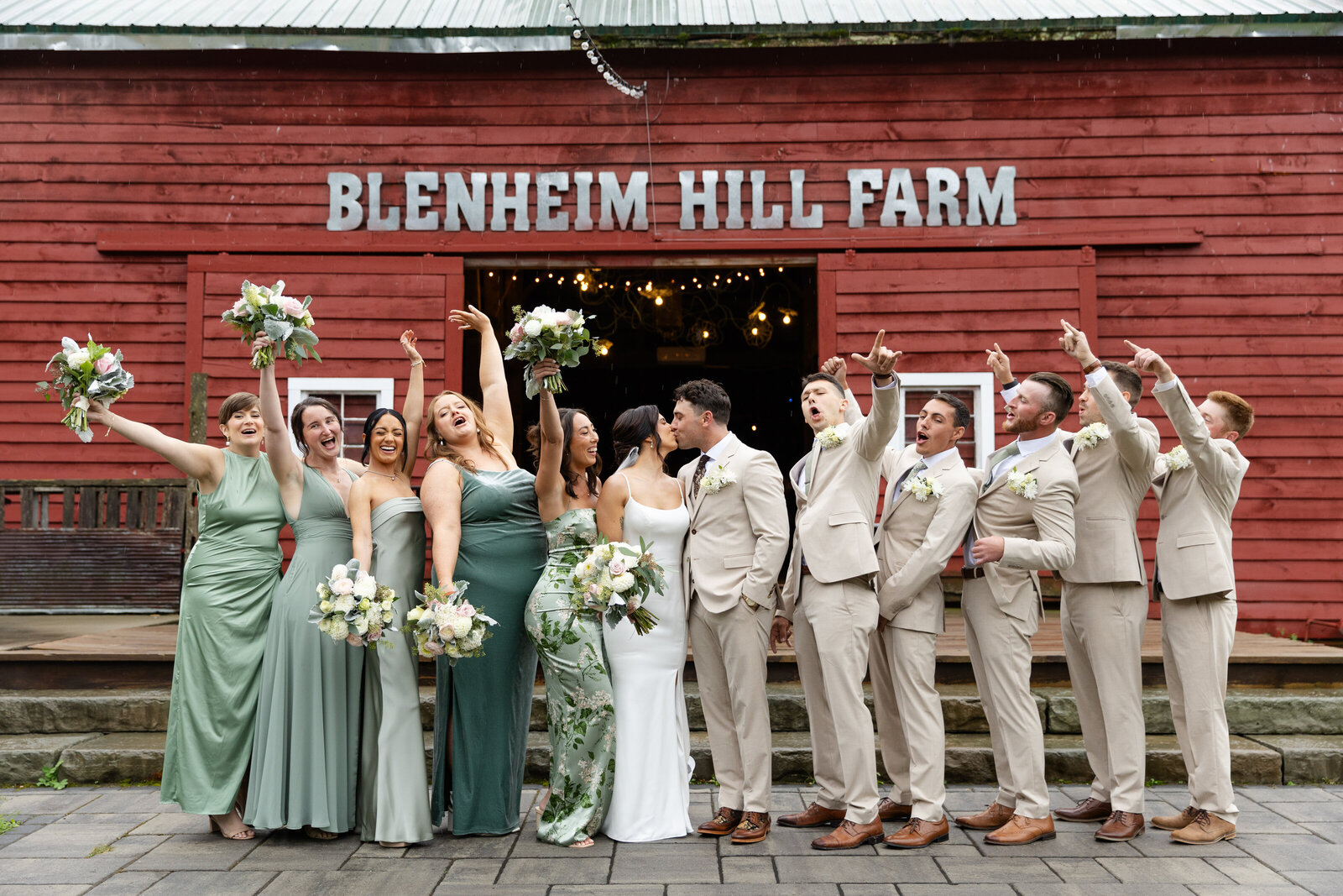 Full bridal party cheers as bride and groom share a kiss in front of a barn