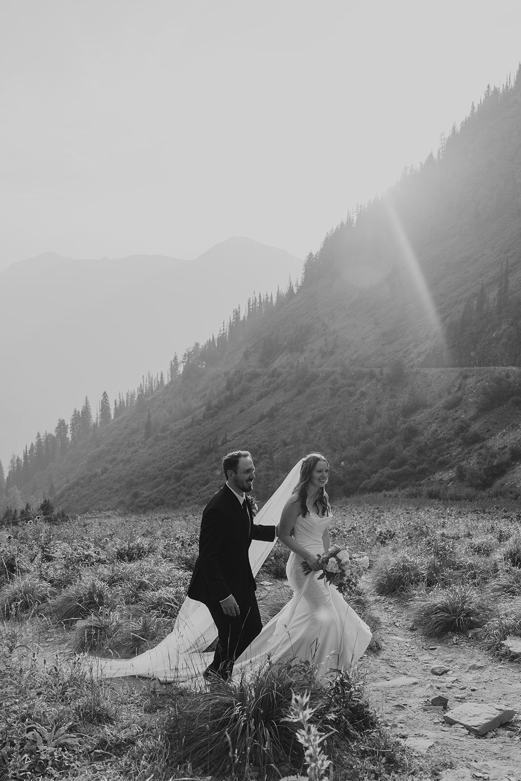 A bride and groom walk through a sunlit mountain valley surrounded by mist and pine-covered slopes during their Glacier National Park elopement, captured in black and white by Sydney Breann Photography.