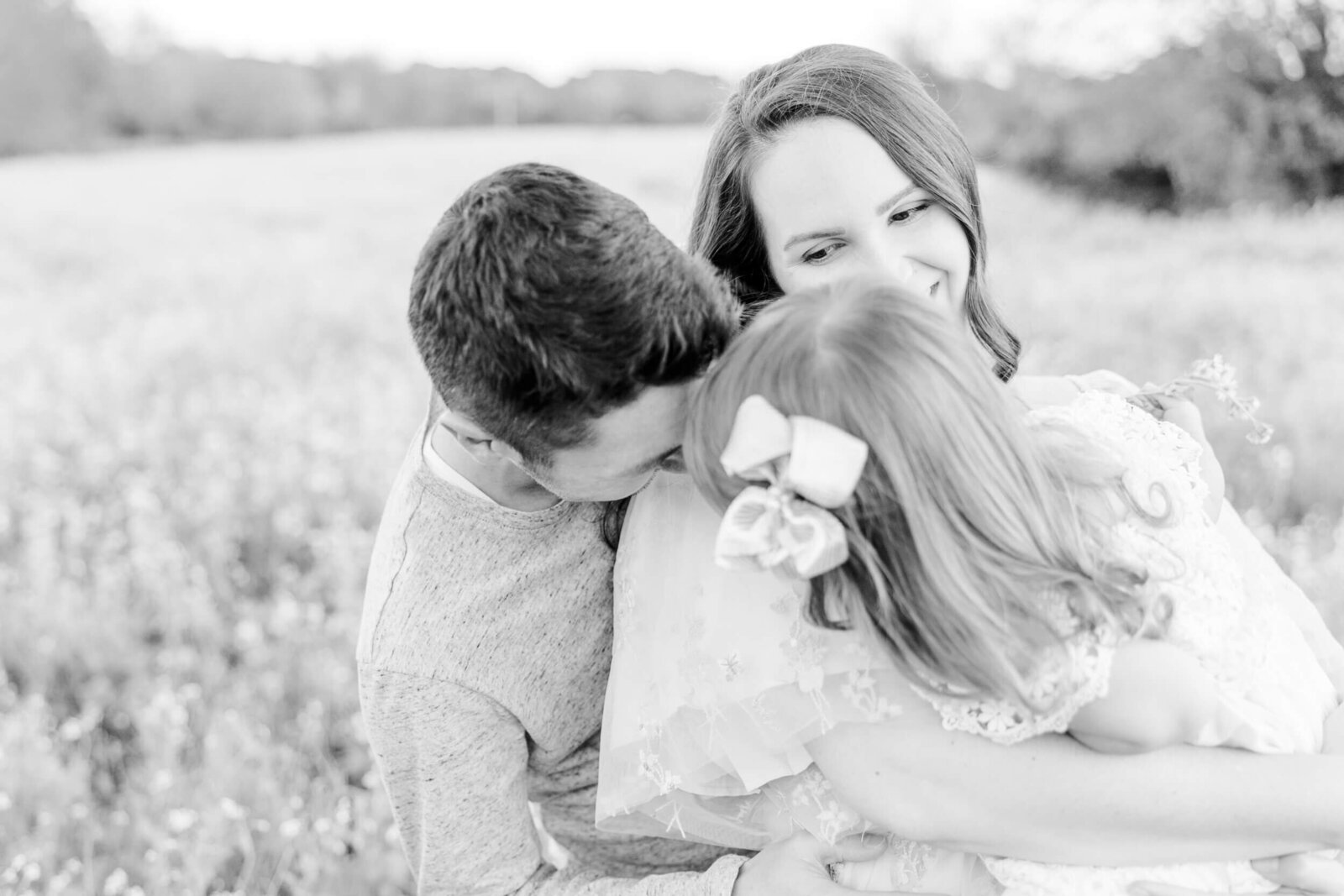 Young girl in mom's arms kisses her dad, who is standing behind her smiling mom