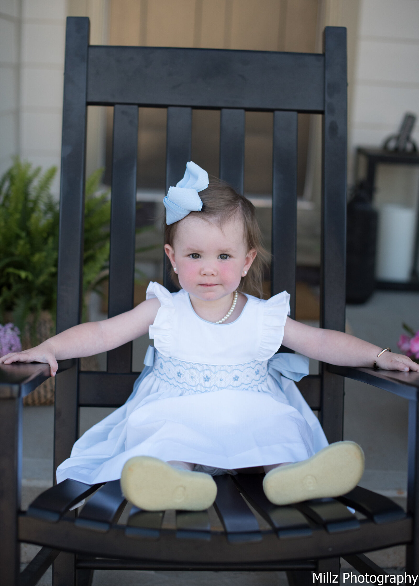 baby girl dressed in a white dress with a bow on her head sitting on a wooden chair photographed by Millz Photography in Greenville, SC
