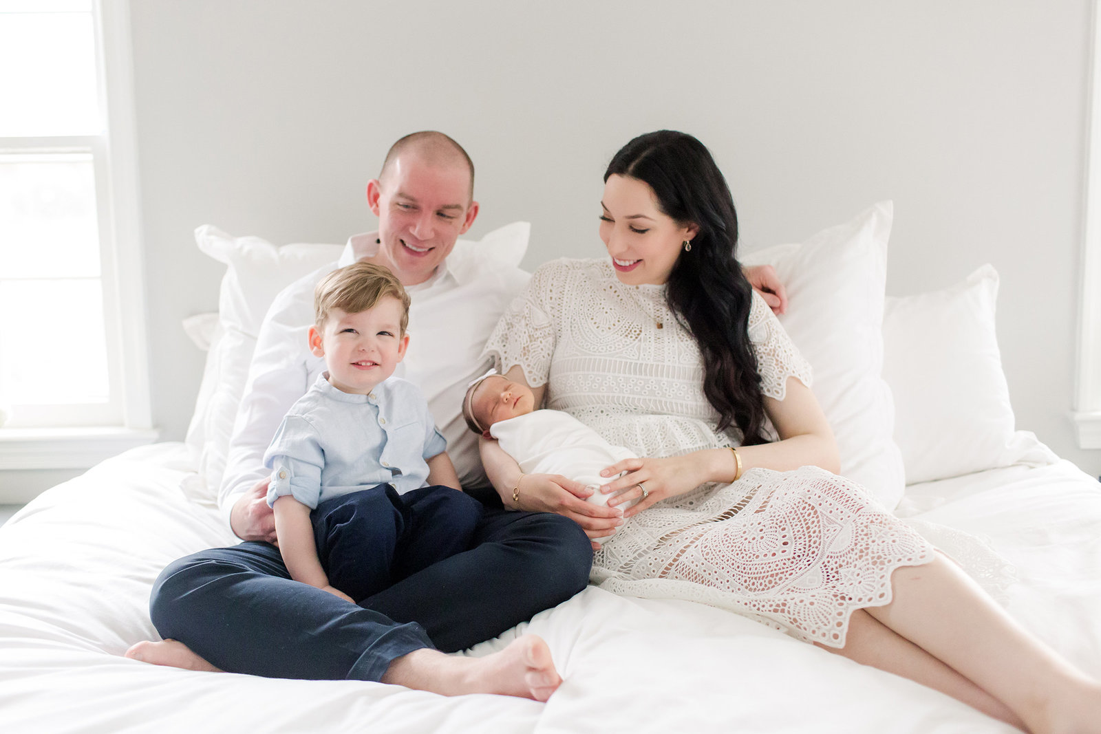 Family sitting on white bbed and smiling at son while holding newborn