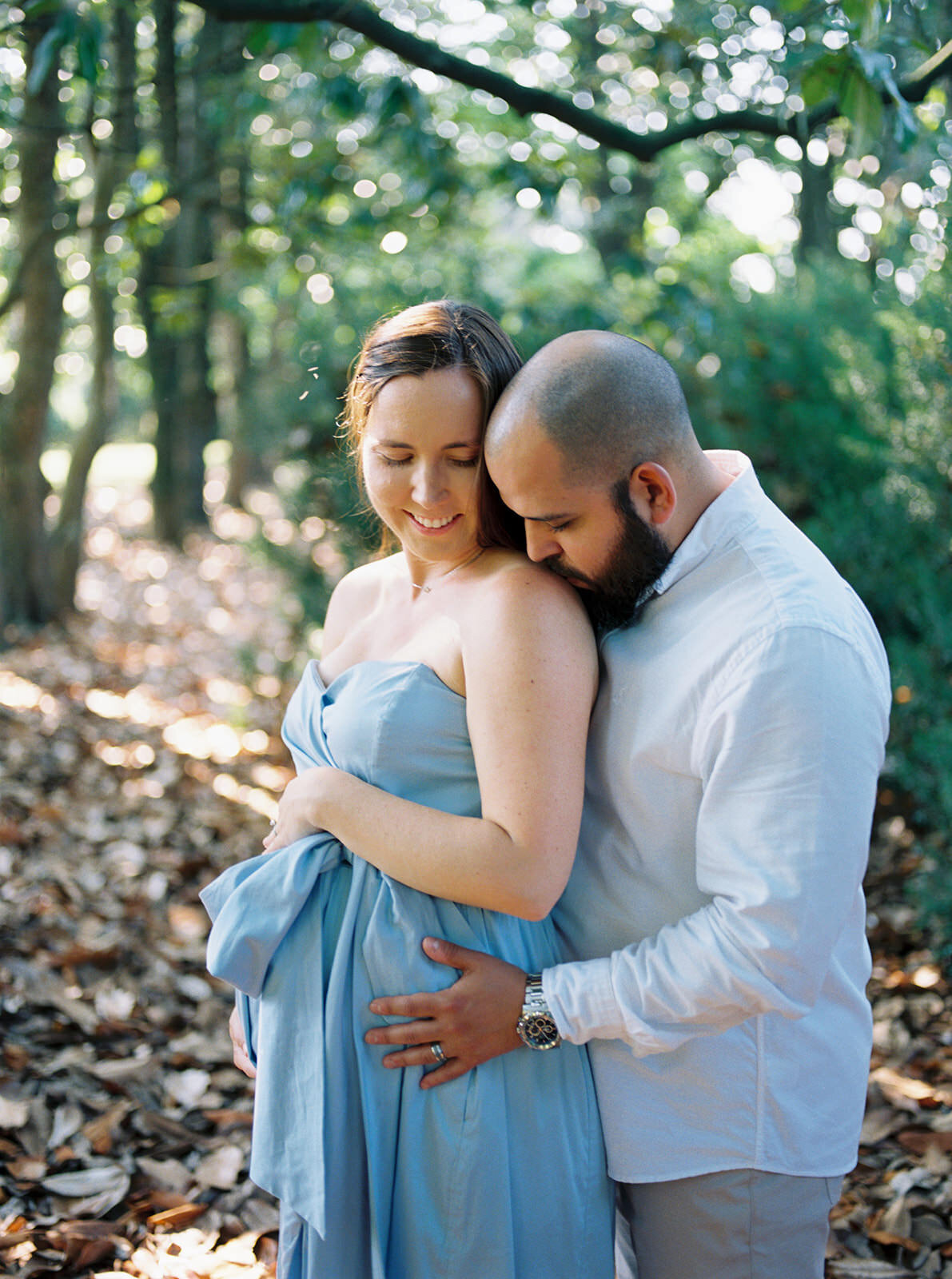 Maternity portrait in a wooded area with soft lighting, capturing a romantic moment between the couple.