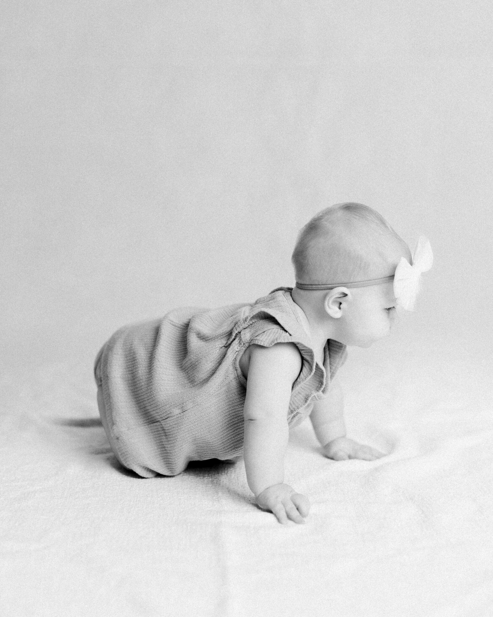 Black and white photo of baby in romper and white bow crawling away in a neutral studio by NH newborn photographer Fieldstone Studio.