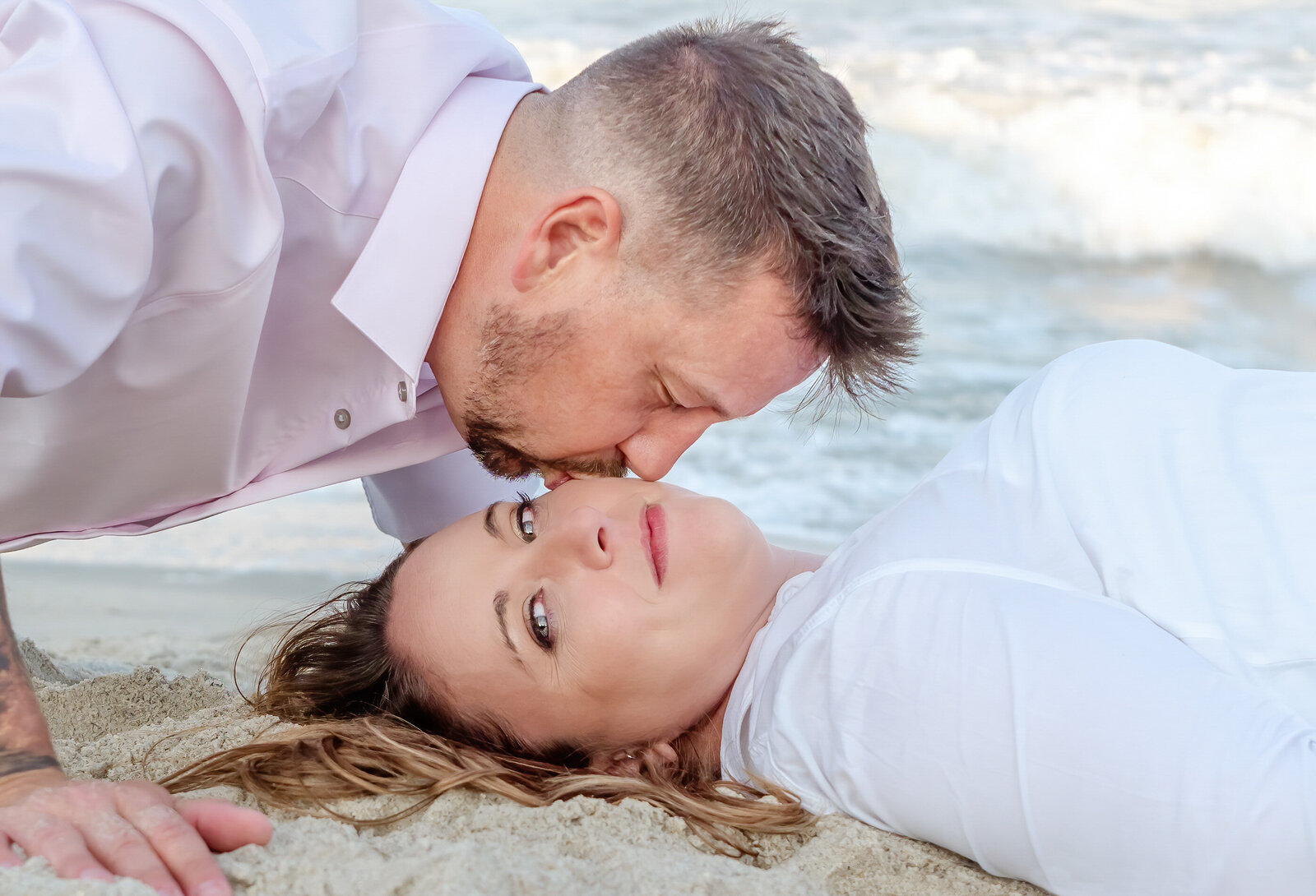 A romantic moment unfolds on the beach as a man gently kisses the forehead of a woman lying in the sand. The waves roll softly in the background, and the couple's relaxed pose captures the intimate, heartfelt style of North Carolina engagement photography. 