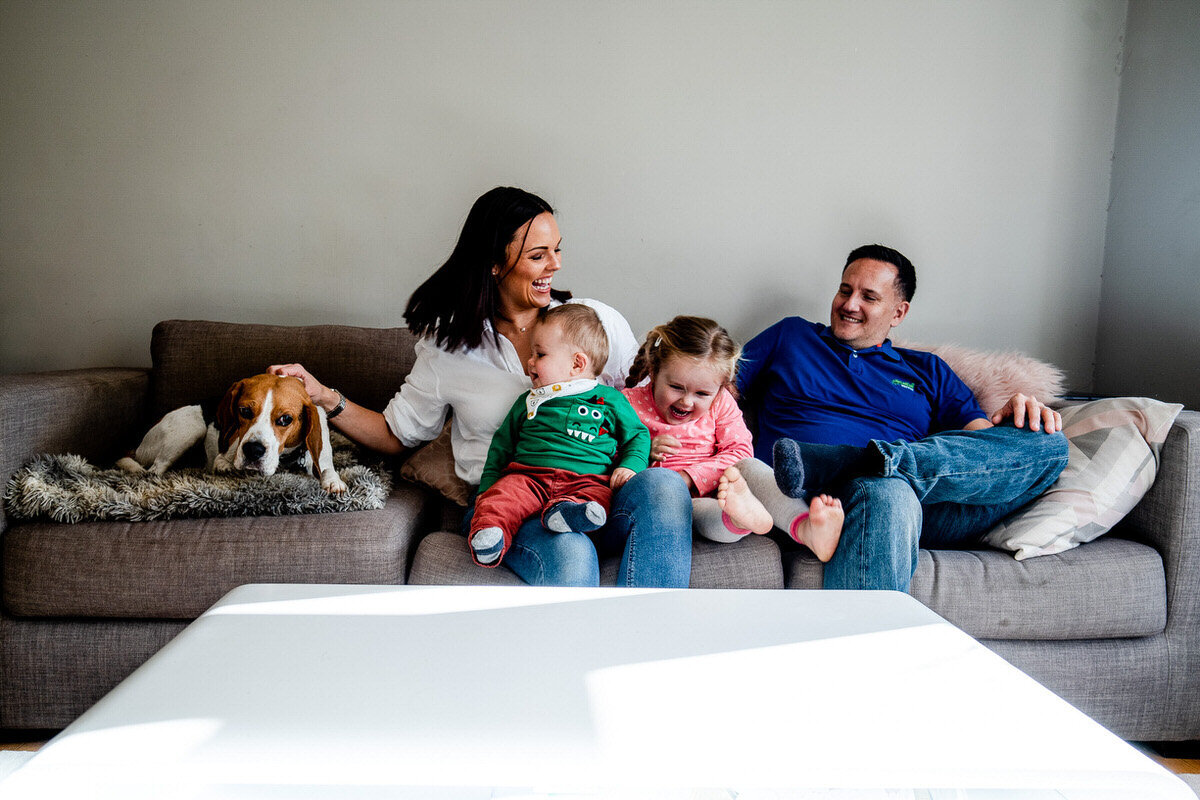 a family sit together on a sofa laughing with one another whilst the dog looks annoyed