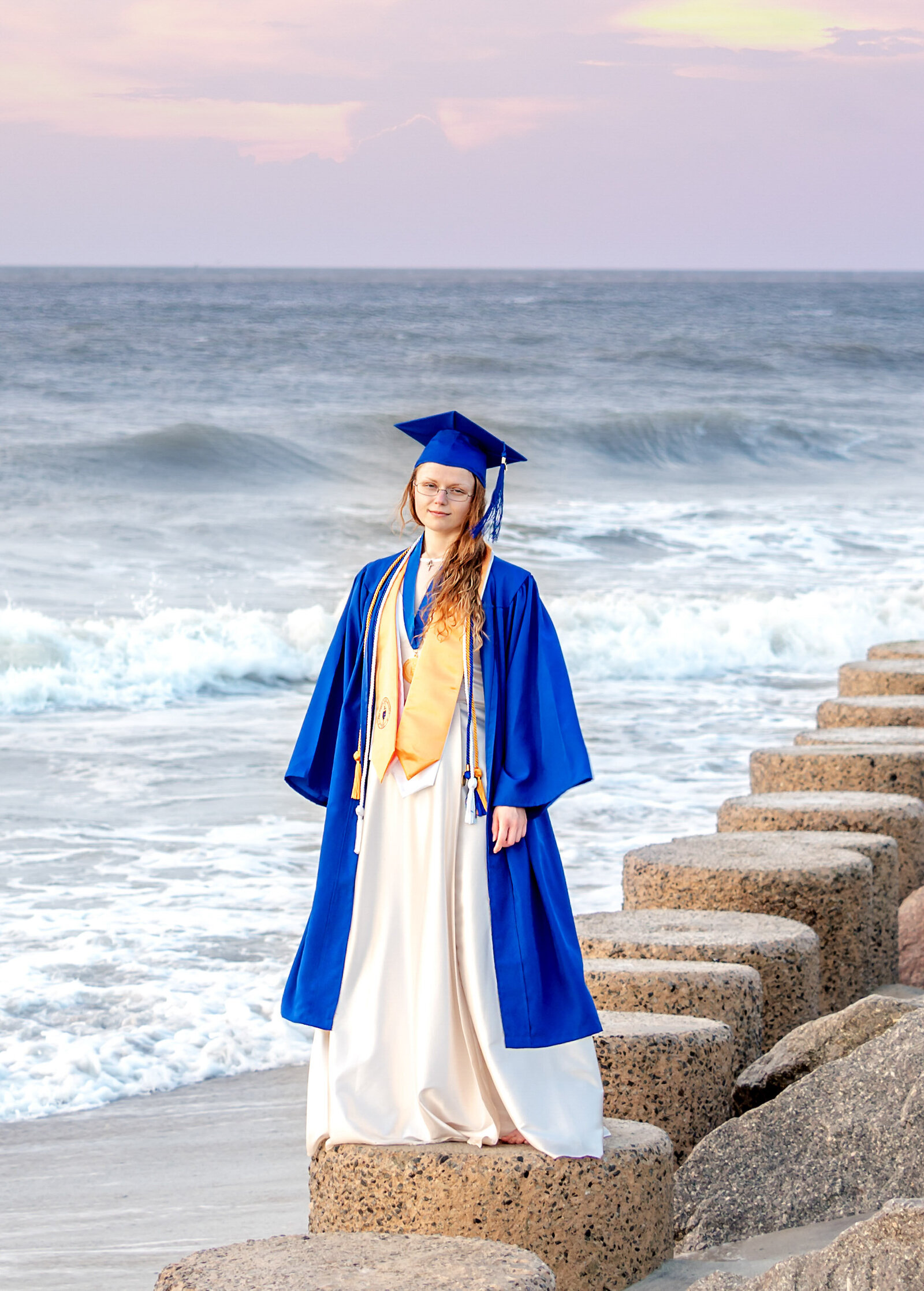 A Cape Fear Community College senior in a blue cap and gown standing on Fort Fisher beach rocks with ocean waves in the background near Wilmington, North Carolina. This striking image captures the celebration of graduation, ideal for artistic senior photography by the sea.