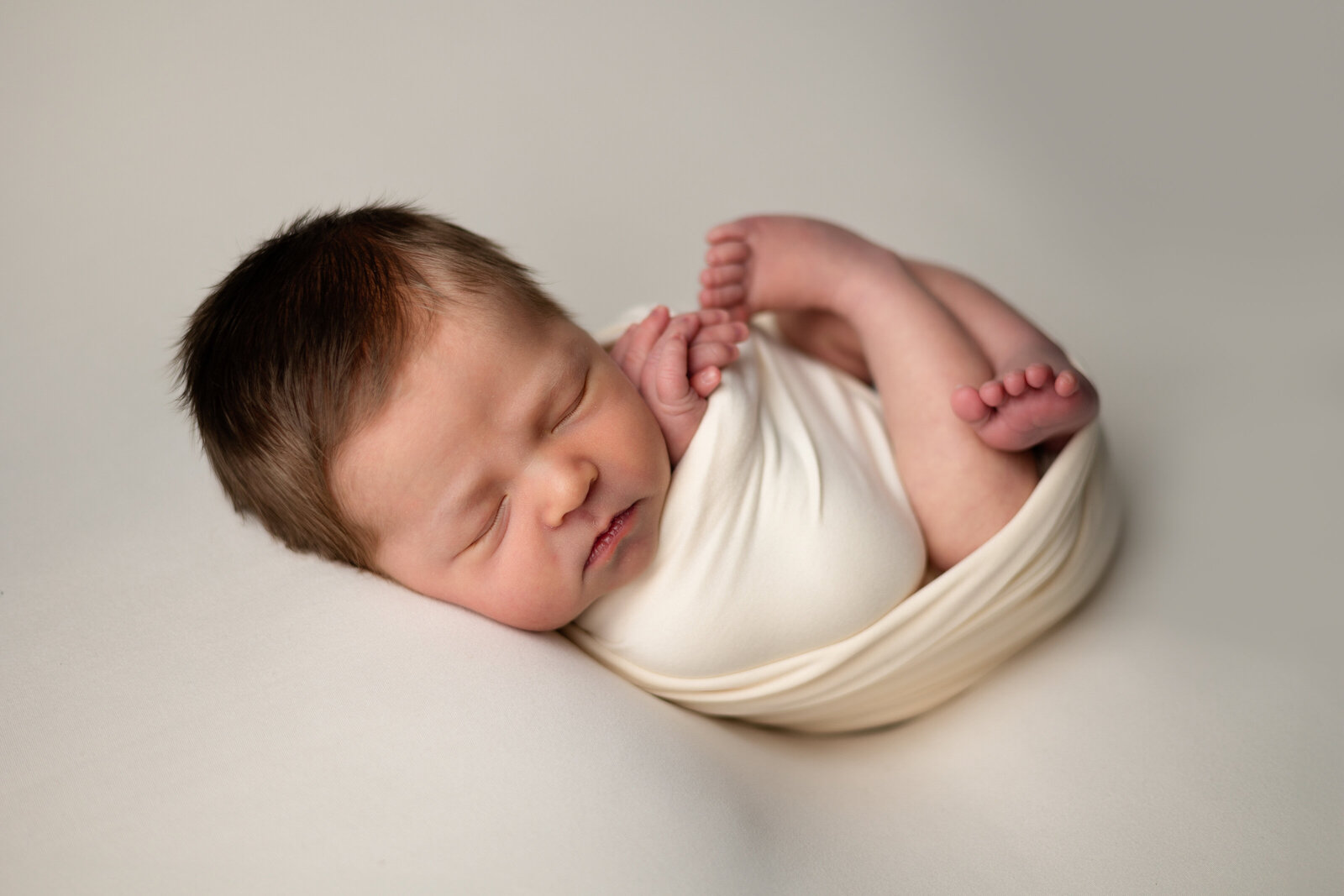 Newborn baby boy swaddled  in a cream wrap and posed on a cream backdrop