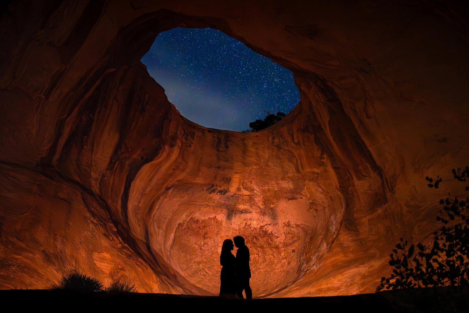 Couple silhouetted underneath Bowtie Arch in Moab under the stars at night during their adventure engagement session.