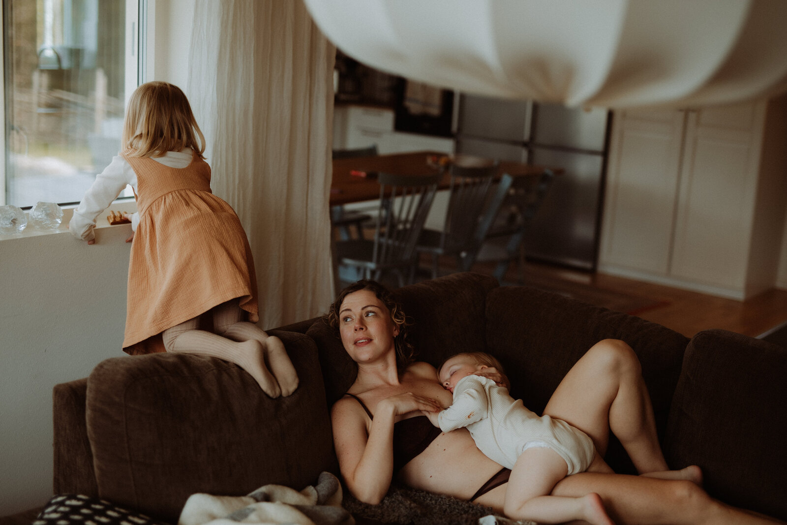 en mamma ligger hemma i sin soffa med sin son på hennes bröst och ammar samtidigt som hennes dotter hoppar i soffan