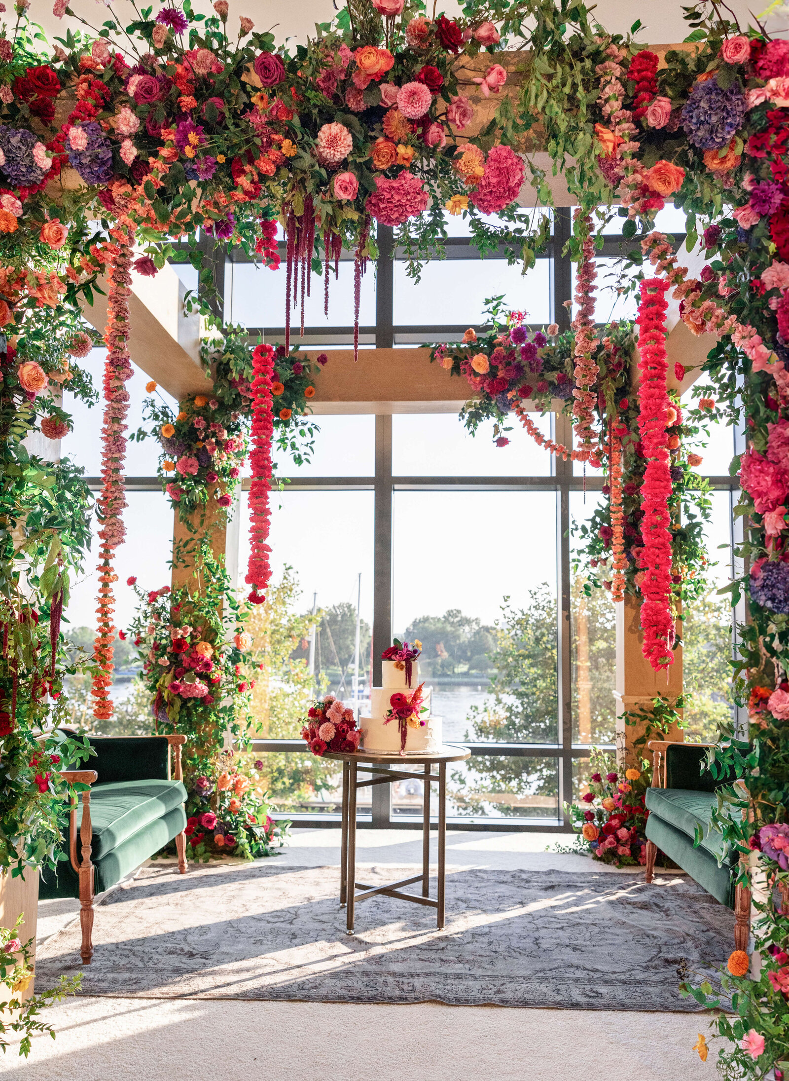 A beautifully decorated indoor wedding altar with vibrant flowers in pink, red, and purple shades. The altar features a small table with a cake, set against large windows overlooking a scenic lake and lush greenery outside.