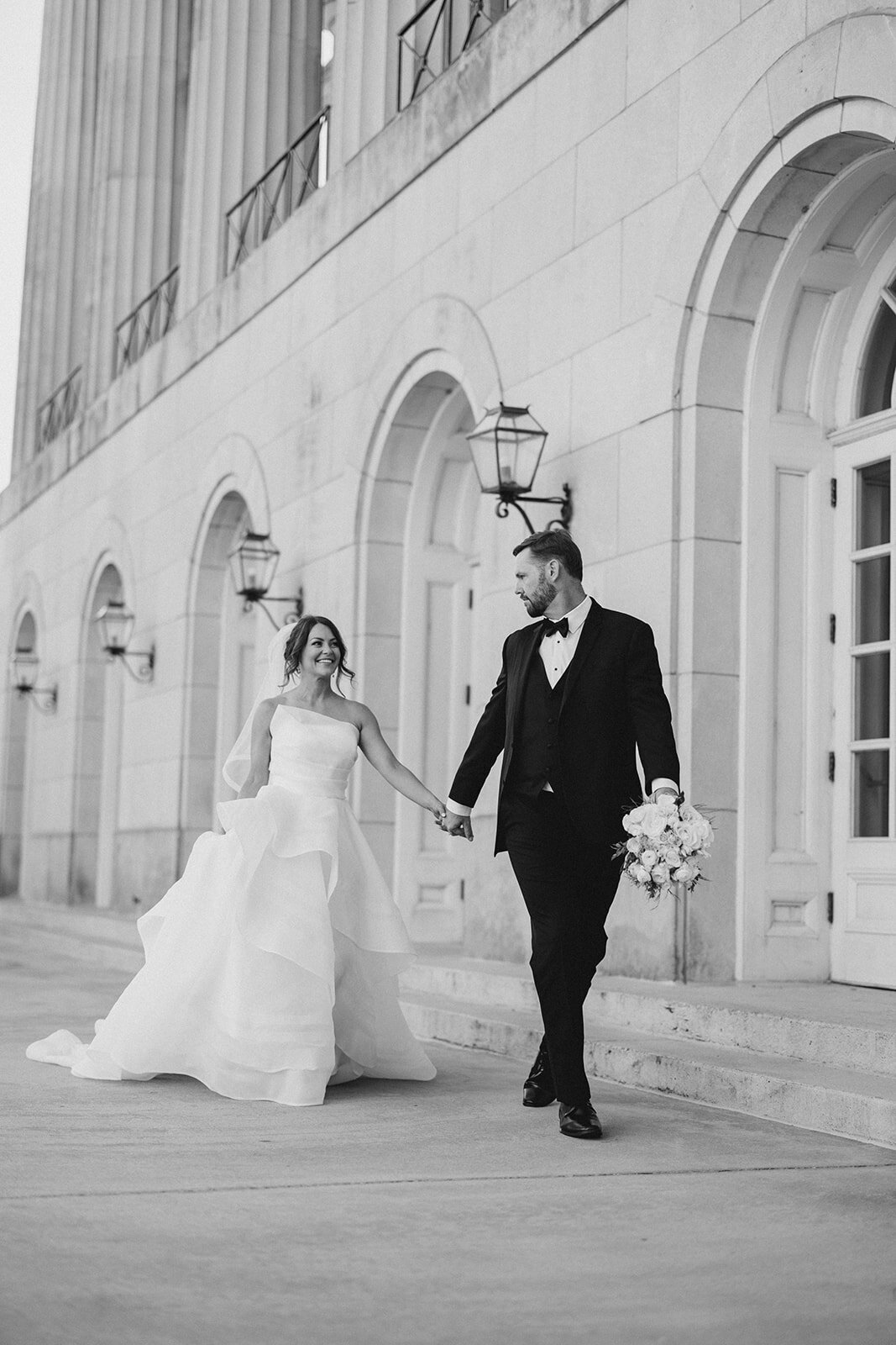 Bride follows groom in black tux in front of European building at destination wedding