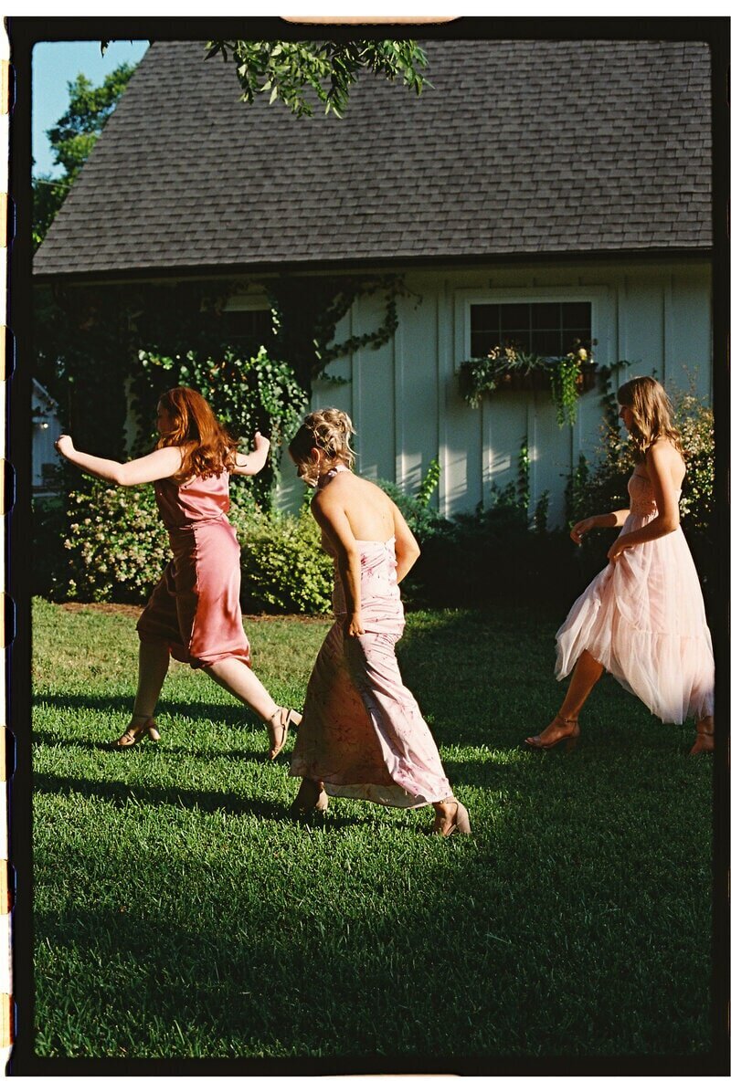 Three women in pink dresses walk on a lawn in front of a white house with a shingle roof. They appear to be joyful and engaged in conversation, with greenery and floral arrangements adorning the house.