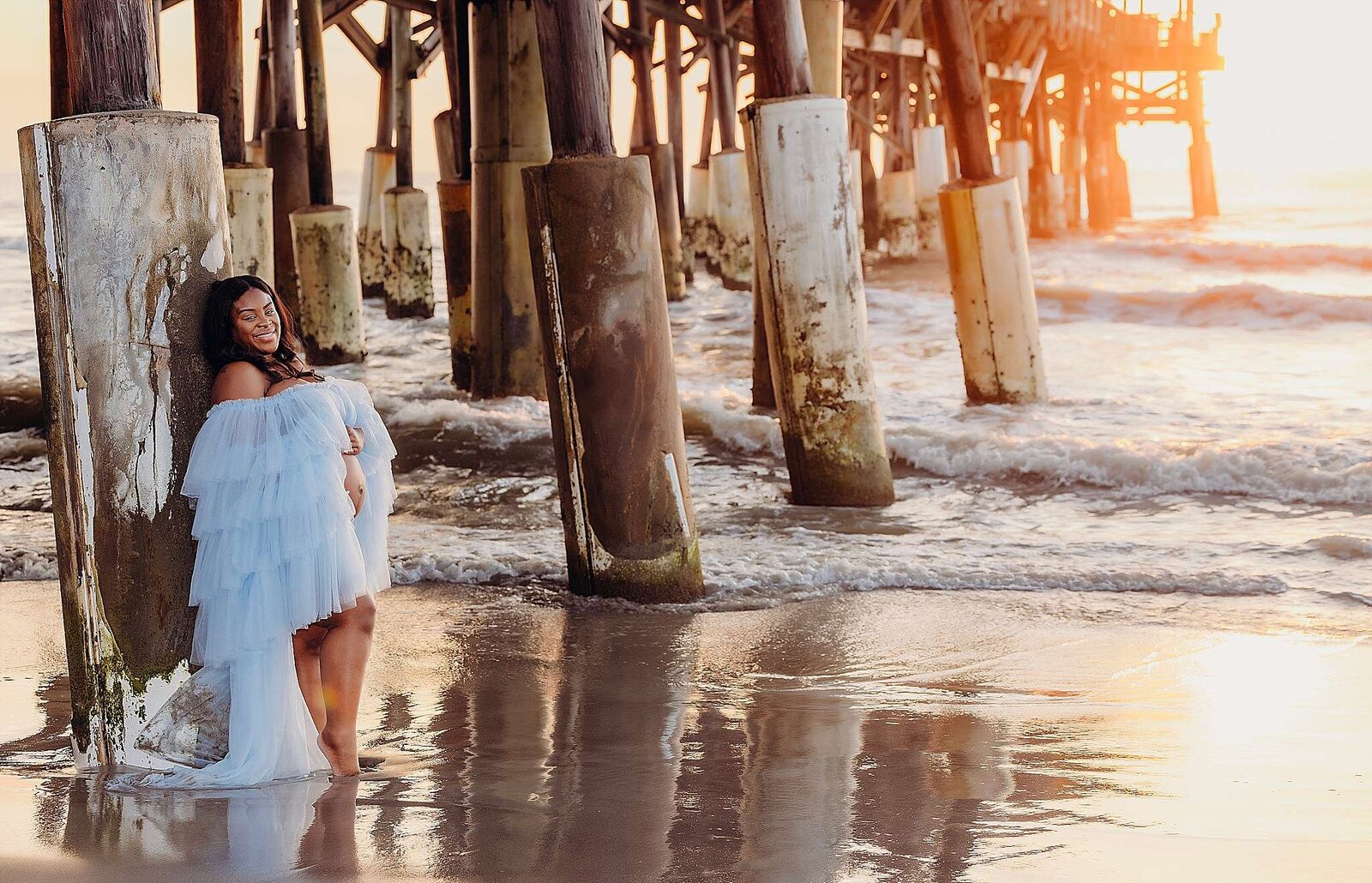 A pregnant woman in a blue dress stands on the beach under a pier, captured by Bia Schaefer Photography in Cocoa Beach, Fl