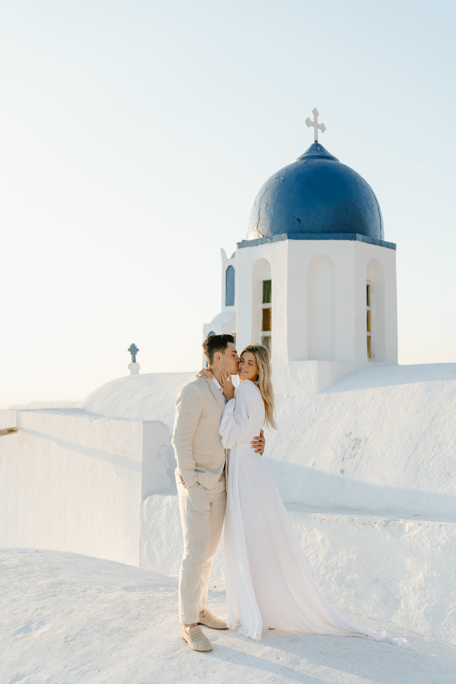 Couple dressed in wedding attire eloping on top of a white dome church on the cliffs of Santorini Greece at Sunset during golden hour.